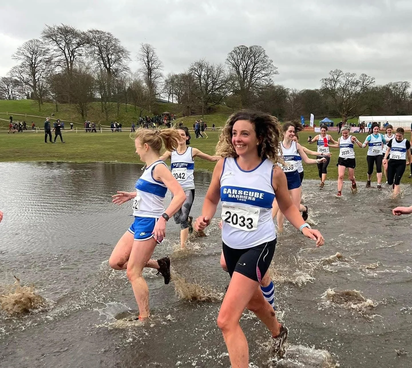 Women participating in a beach race, running through water with onlookers and trees in the background.