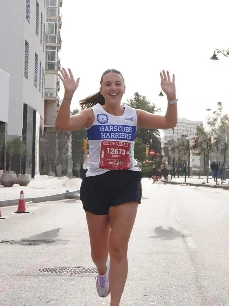 Female runner participating in a race, smiling, wearing a Garscure Harriers tank top and bib number 872, running on a tree-lined road.