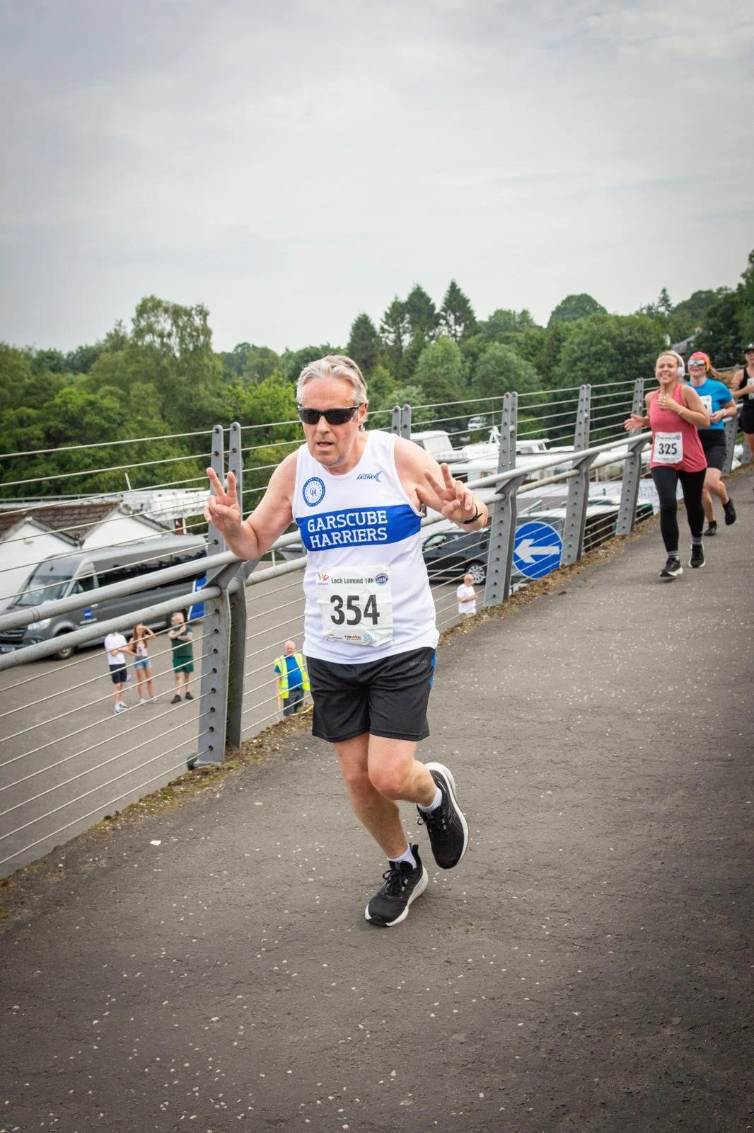 A man in running attire with bib number 354 participating in a race, making peace signs with both hands while running on a paved path beside a metal railing, with several other runners behind him, trees, and parked cars in the background.