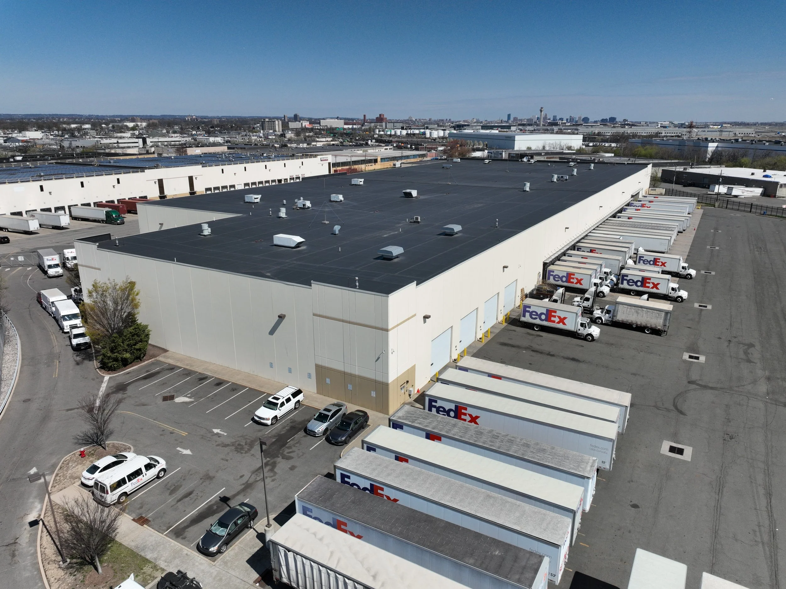 A large warehouse with a black flat roof surrounded by parking lot and delivery trucks, including multiple FedEx trucks. The background features a city skyline with tall buildings under a clear blue sky.