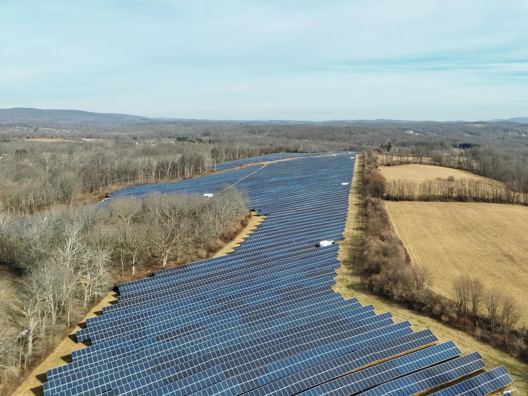 A large solar farm with rows of solar panels stretching across a wide field surrounded by trees and farmland under a partly cloudy sky.