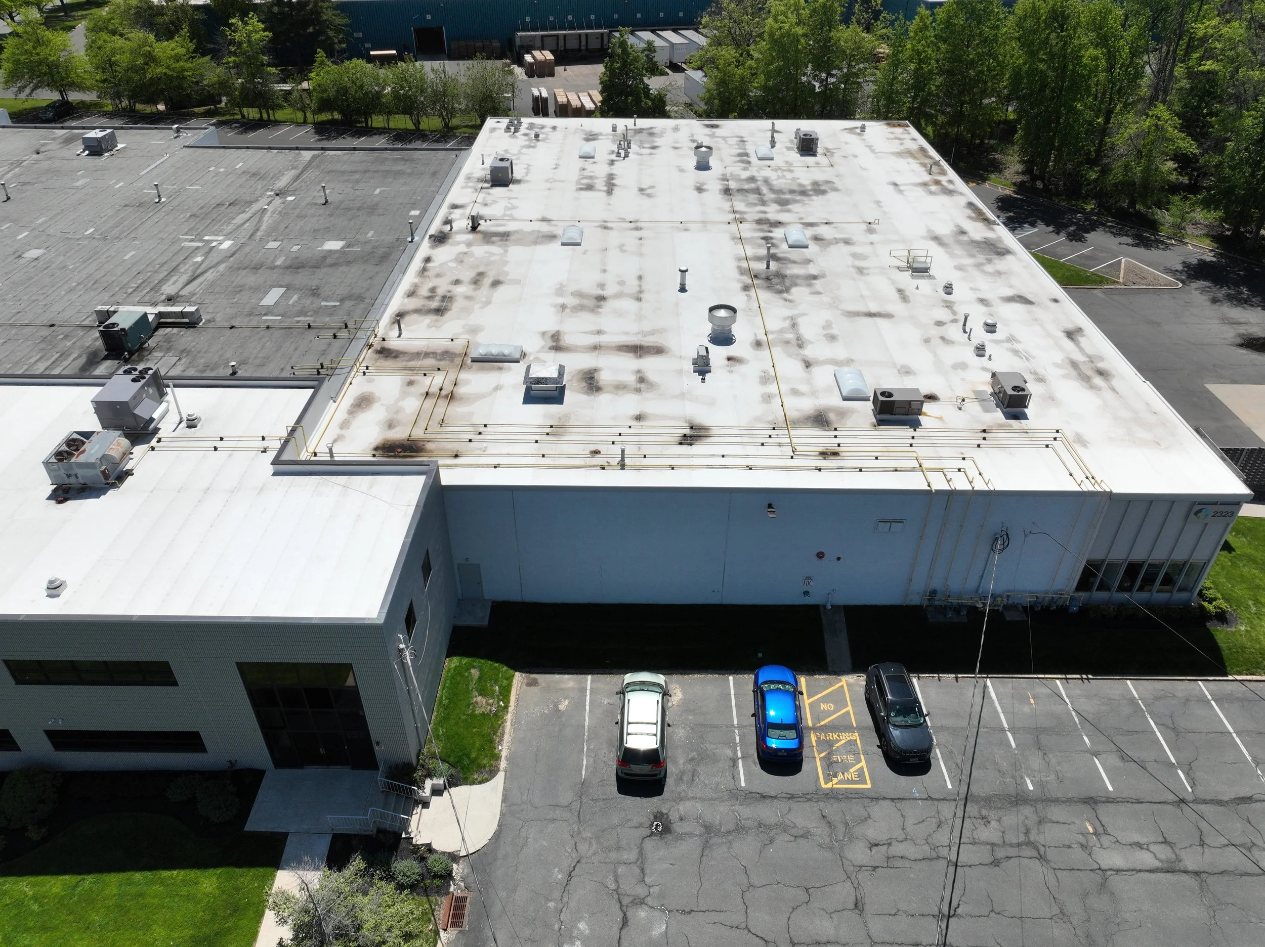 An aerial view of a commercial building's flat roof with HVAC units and vents, surrounded by trees, parking lot, and light traffic.
