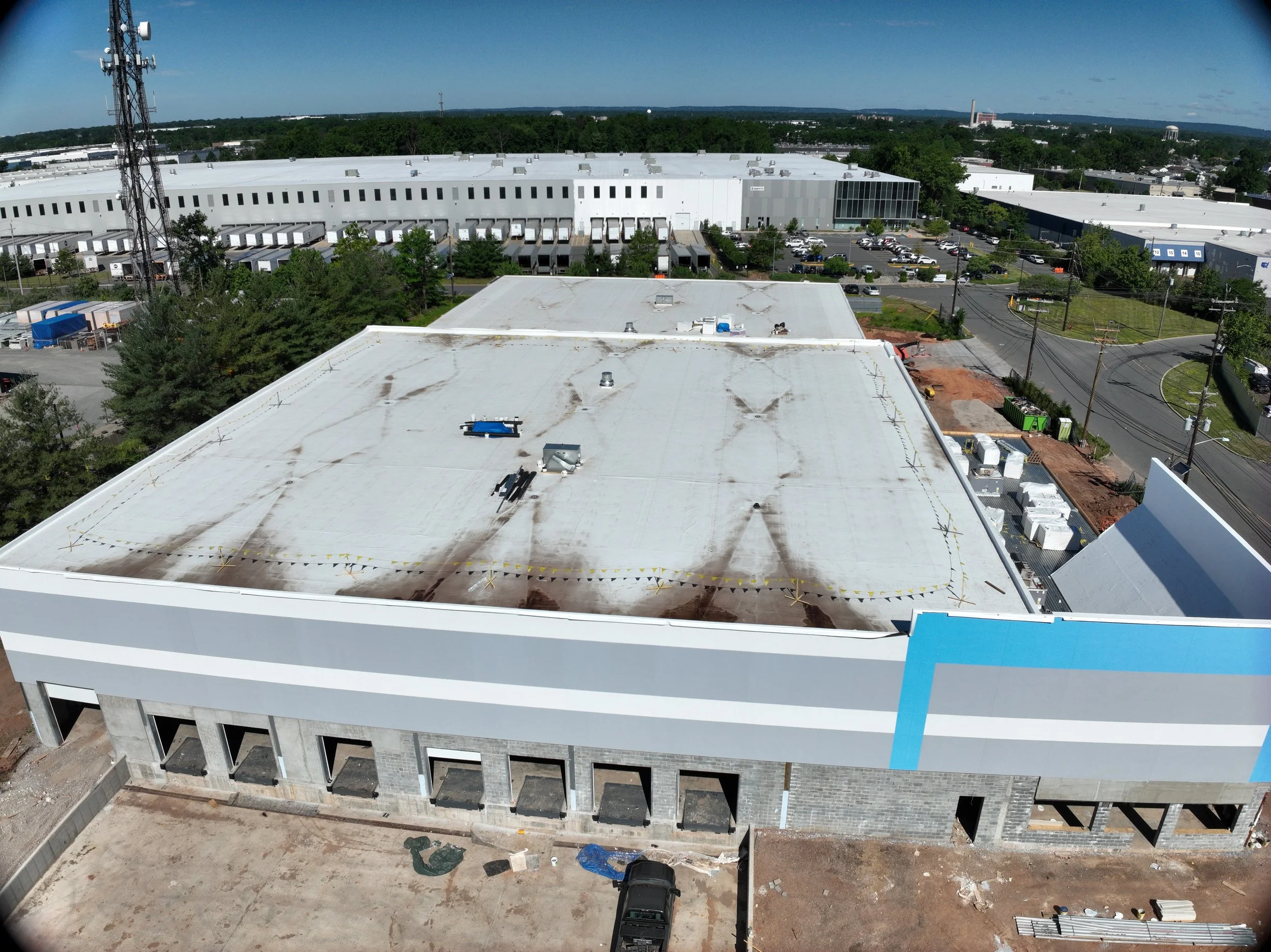 Aerial view of a large industrial or warehouse building under construction with a parking lot filled with cars and trucks, surrounded by trees and power lines.
