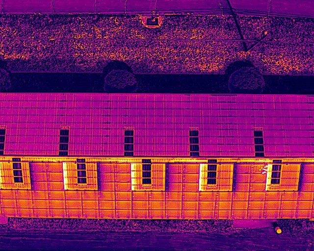 Aerial view of a long building with a sloped roof, showing solar panels on one side and warm-colored lights illuminating the other side at night.