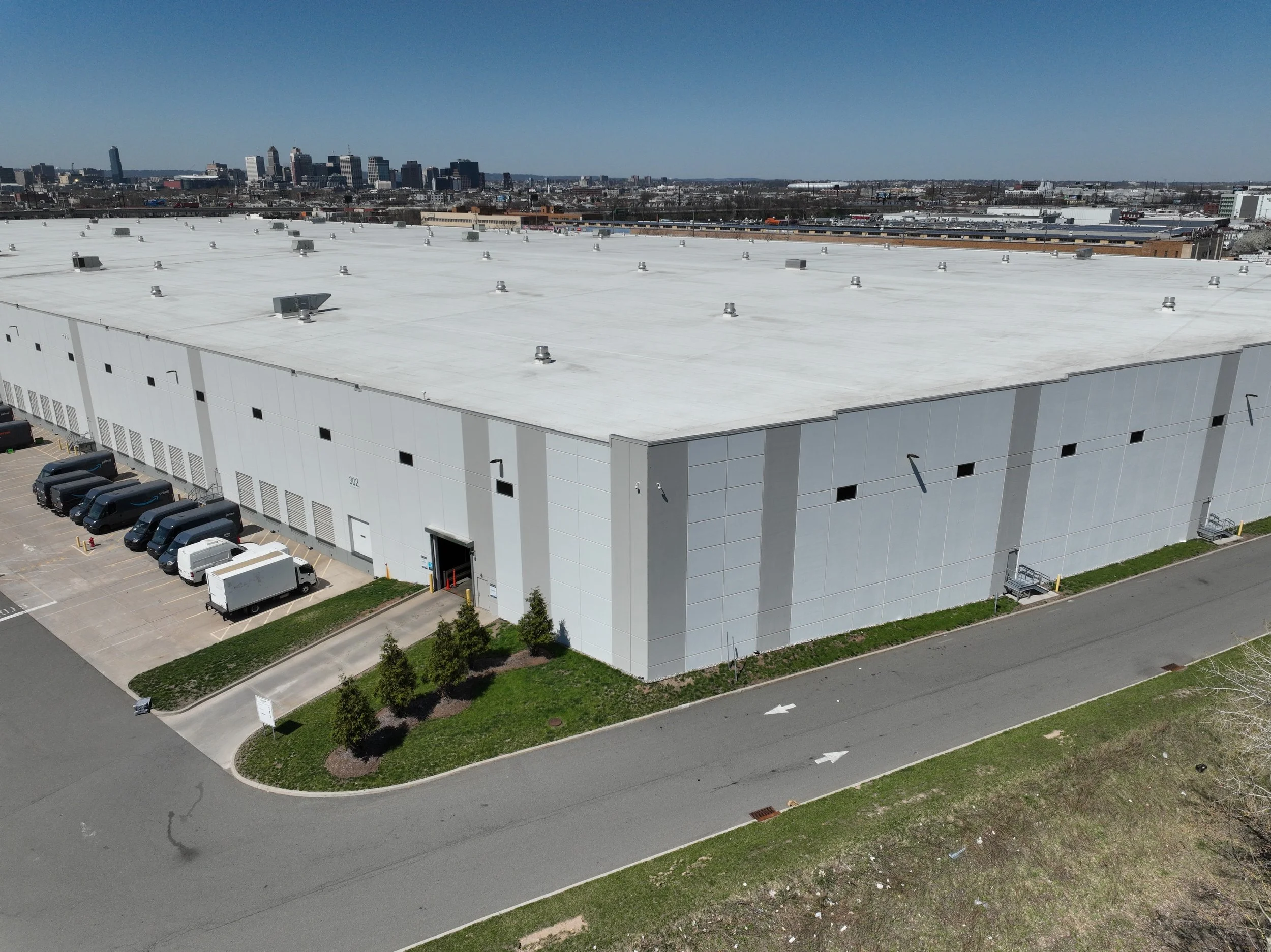 An industrial building with a white flat roof, numerous rooftop ventilation units, and a parking lot with several vans and trucks, set against a city skyline in the distance.