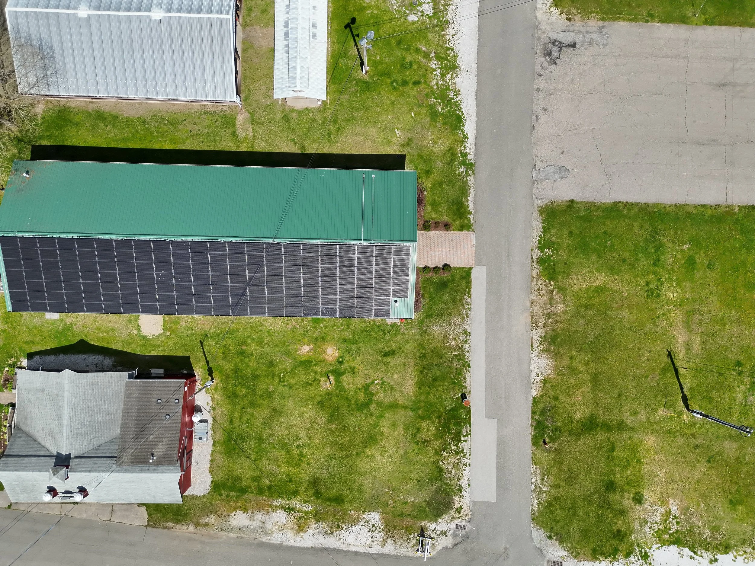 An aerial view of a residential area showing a house with a green metal roof, a satellite dish, a chimney, and solar panels. Surrounding the house are patches of grass, a driveway, and sidewalks. There are streetlights and some trees visible.