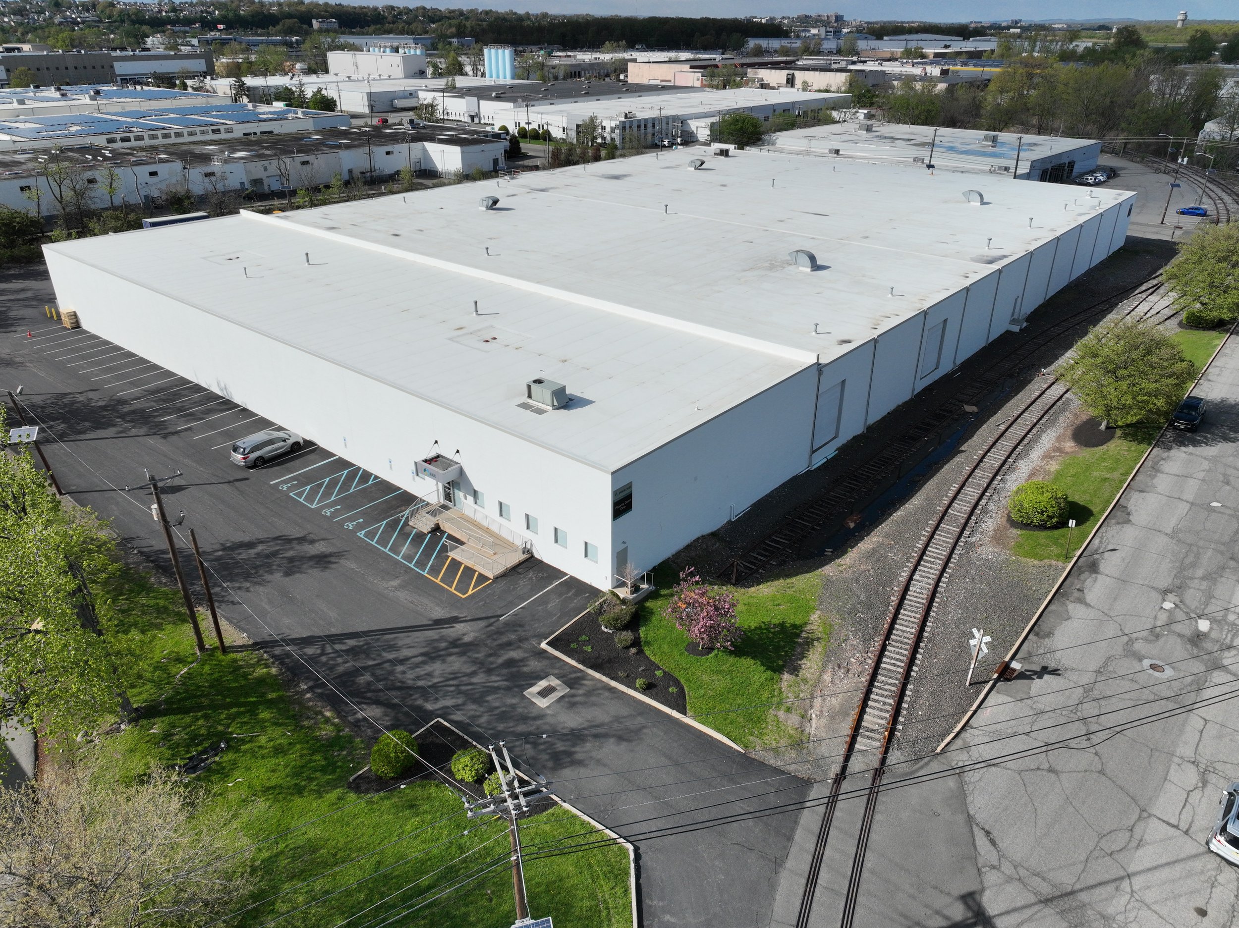 An aerial view of a large white industrial or warehouse building on a sunny day, with adjacent parking lot, surrounding roadways, railway tracks, and some trees along the edges.