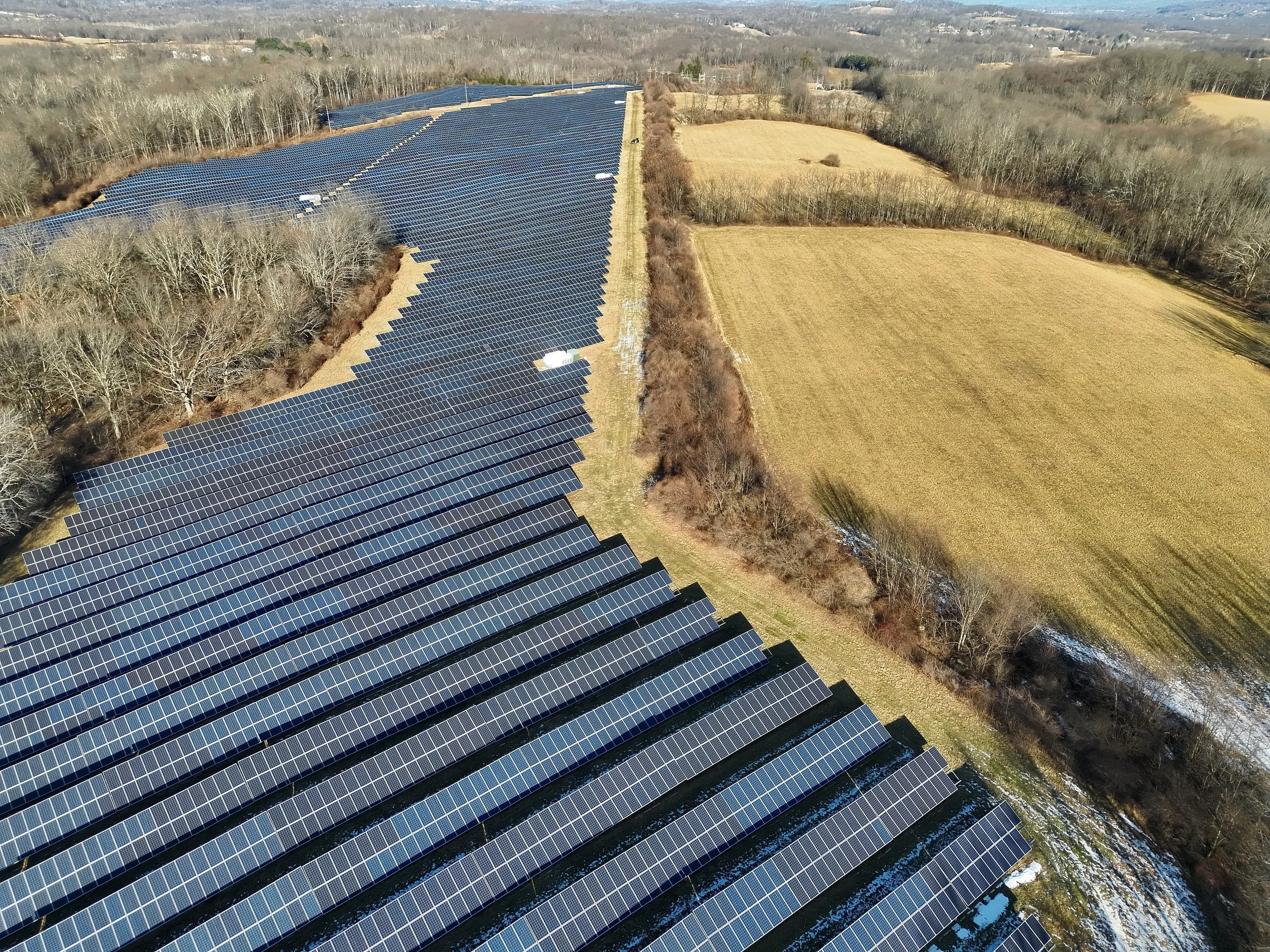 An aerial view of a large solar panel farm situated in a rural area with open fields and trees surrounding it.