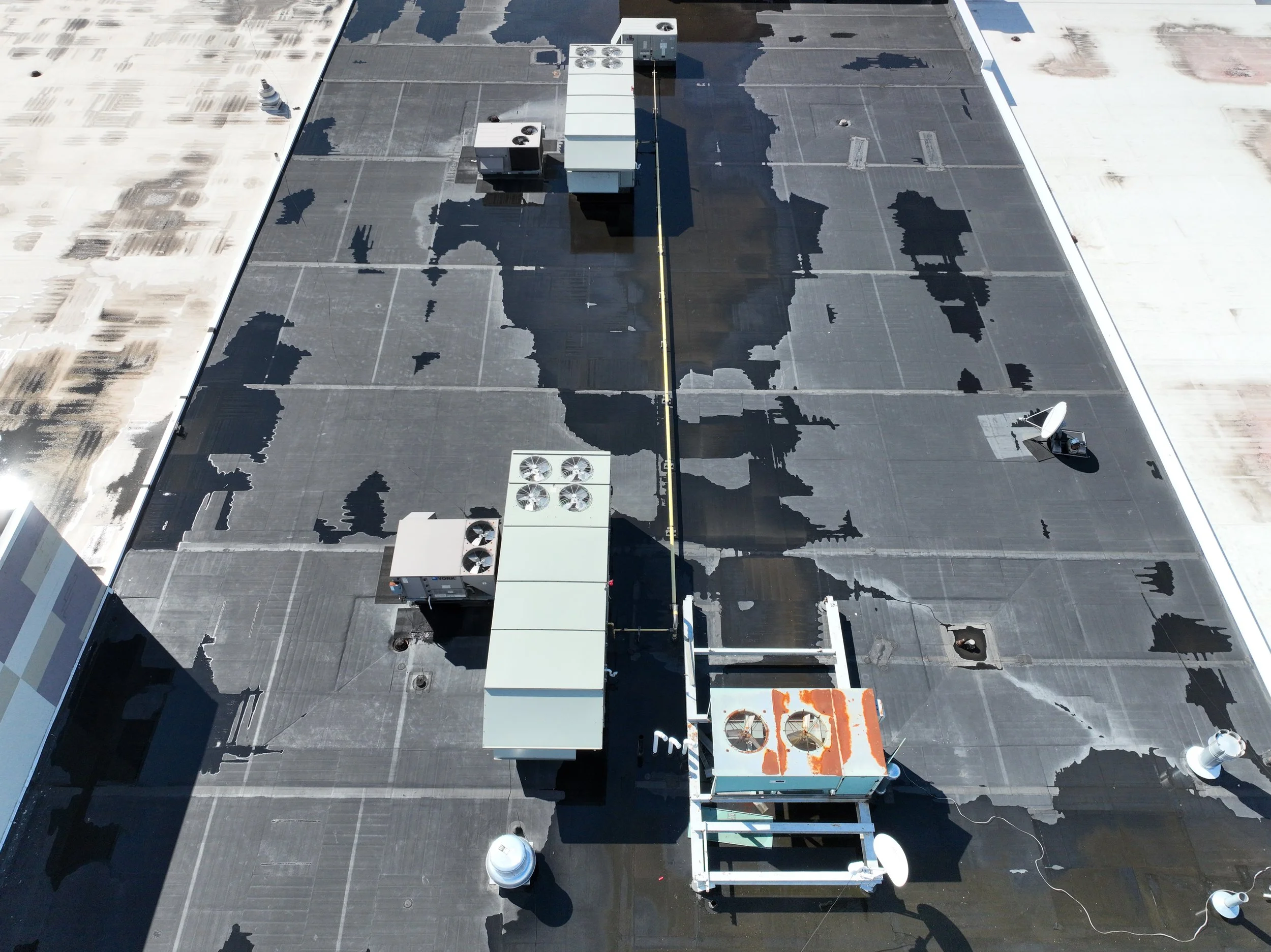 A flat rooftop with black roofing material, HVAC units, and vents, some areas showing patches of weathered or damaged material, and a satellite dish.