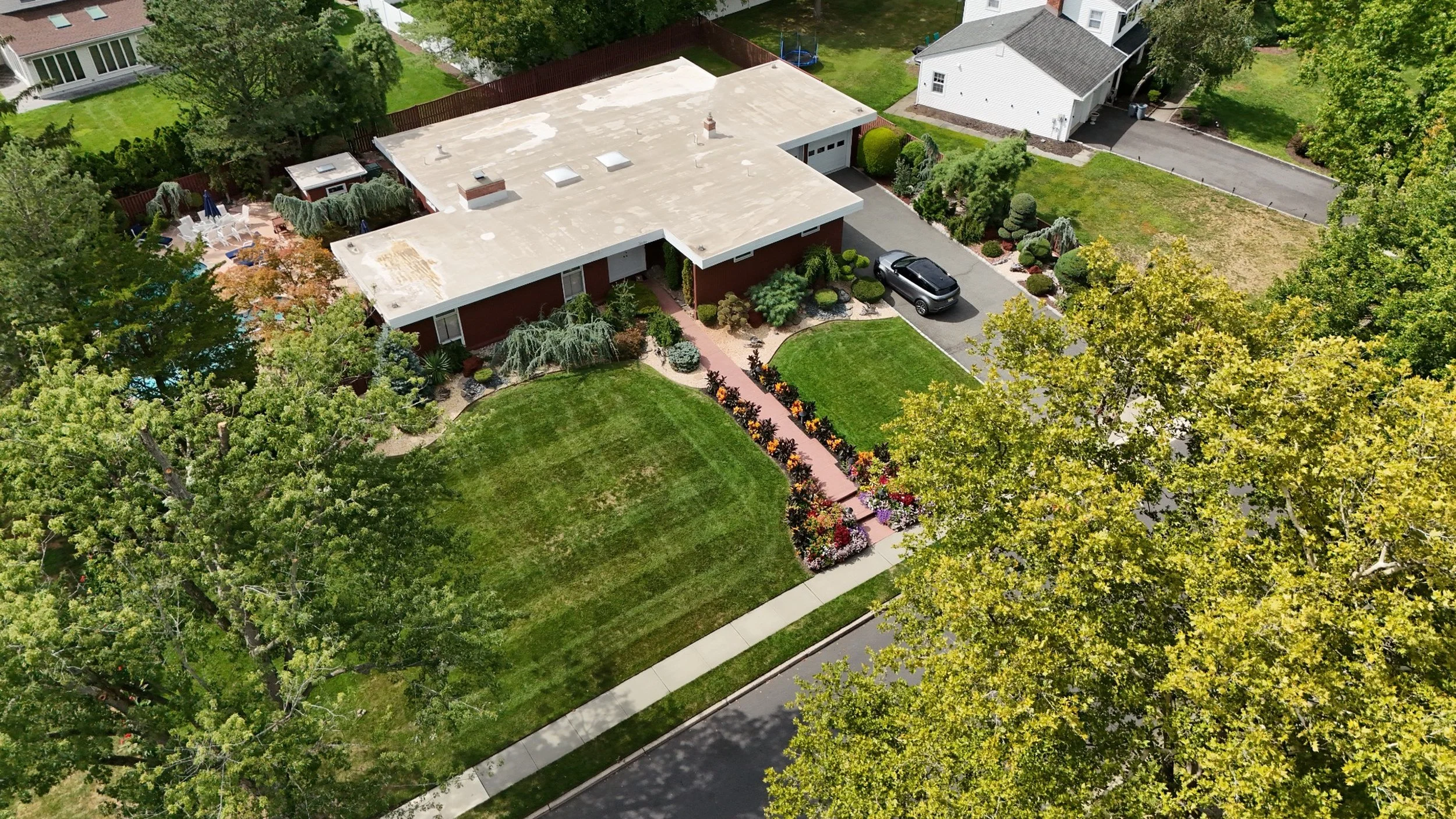 Aerial view of a residential house with a flat roof, surrounded by a well-maintained lawn, colorful flower beds, and trees. There is a driveway with a parked black car and a backyard with outdoor furniture and a swimming pool.