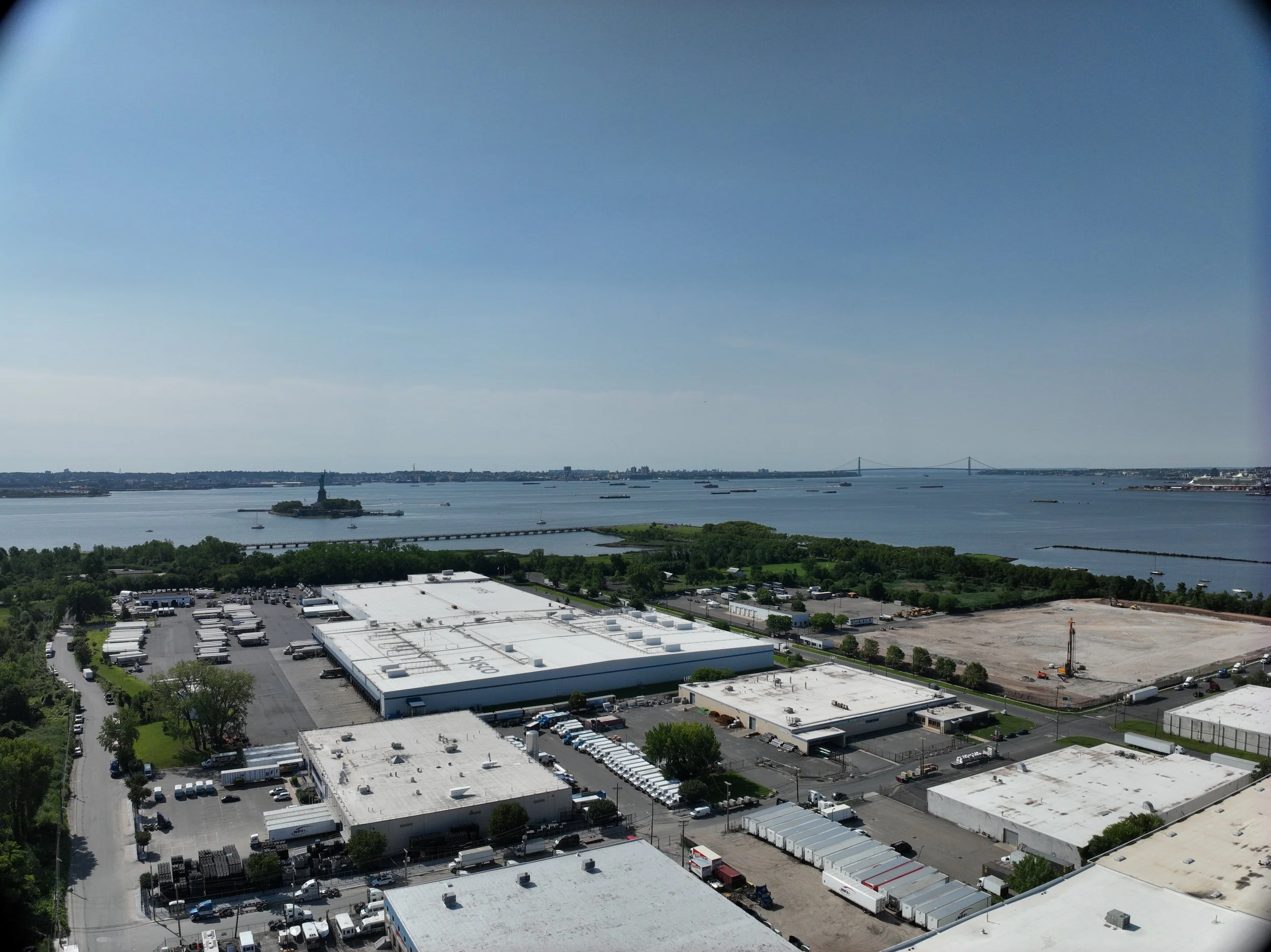 Aerial view of a cityscape with a body of water, the Statue of Liberty on an island, and a distant suspension bridge under a clear blue sky.