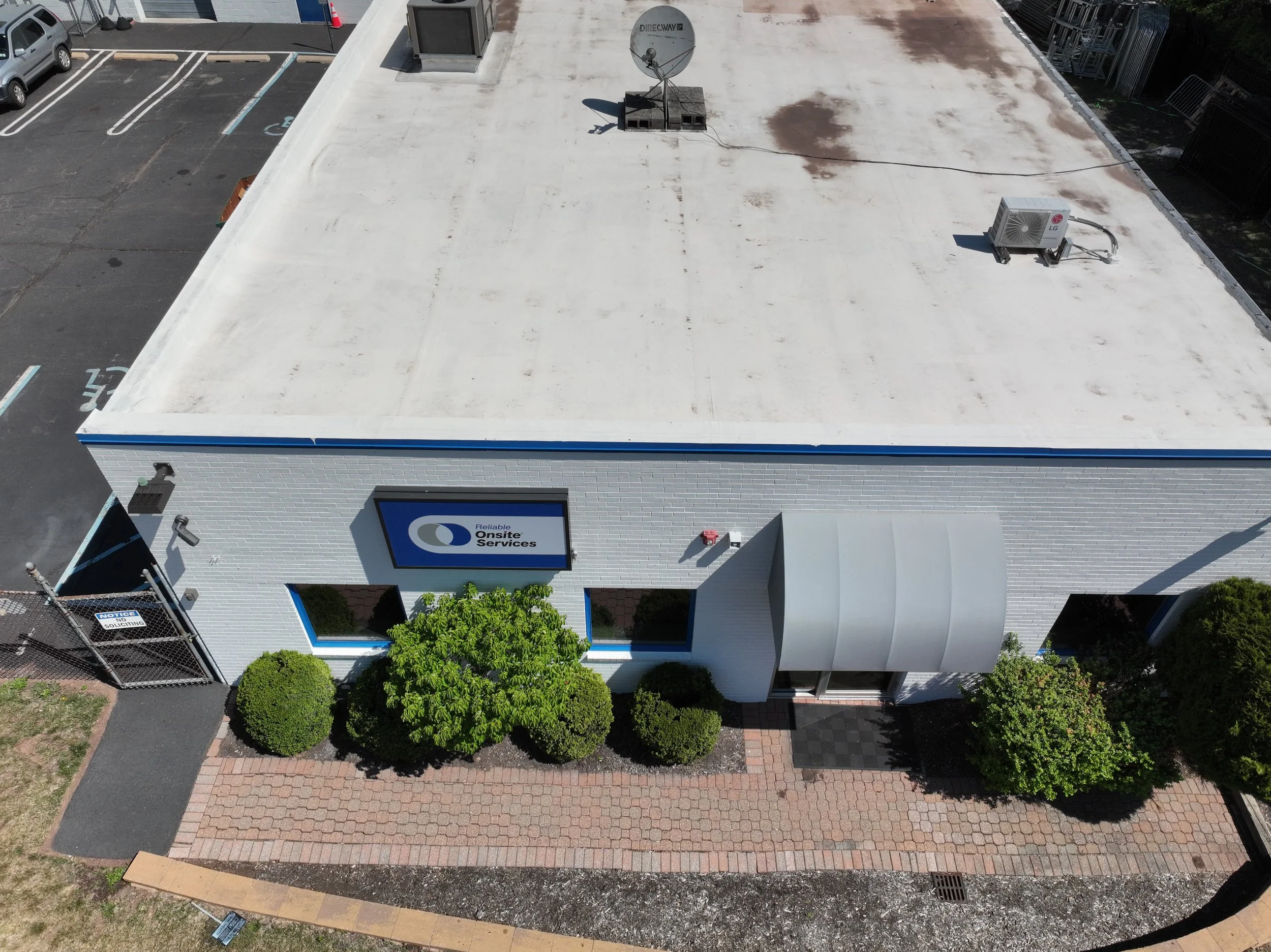 An aerial view of a commercial building with a white flat roof, air conditioning units, and a satellite dish. The building has a sign that says "Reliable Onsite Services." Adjacent parking lot with marked parking spaces, some cars, and a handicap parking spot. Surrounding areas include trees, a brick-paved sidewalk, and a chain-link fence.