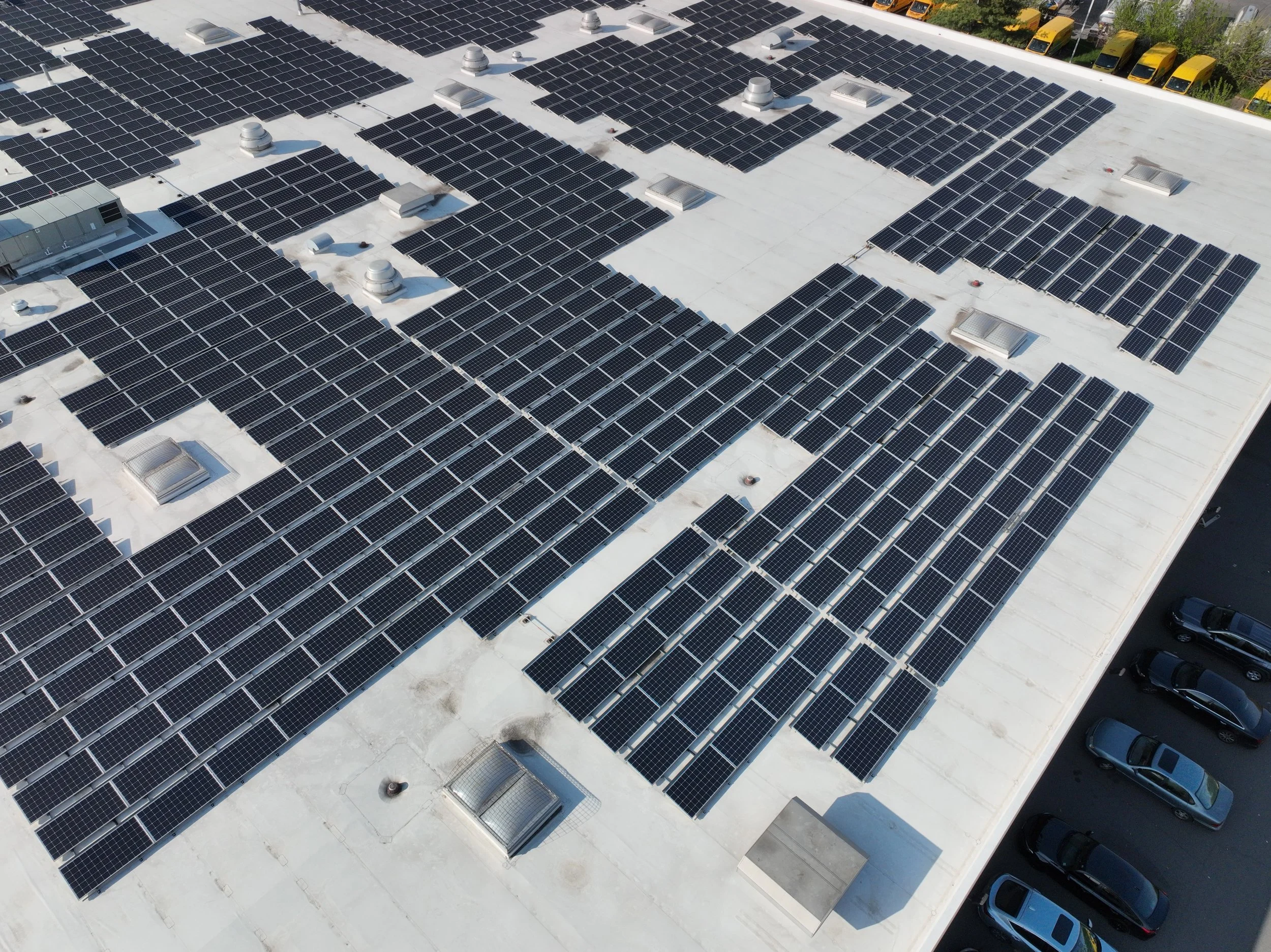 Aerial view of a flat rooftop covered with multiple rows of black solar panels, with some ventilation units and air conditioning units interspersed among the panels. Several cars are parked adjacent to the building.