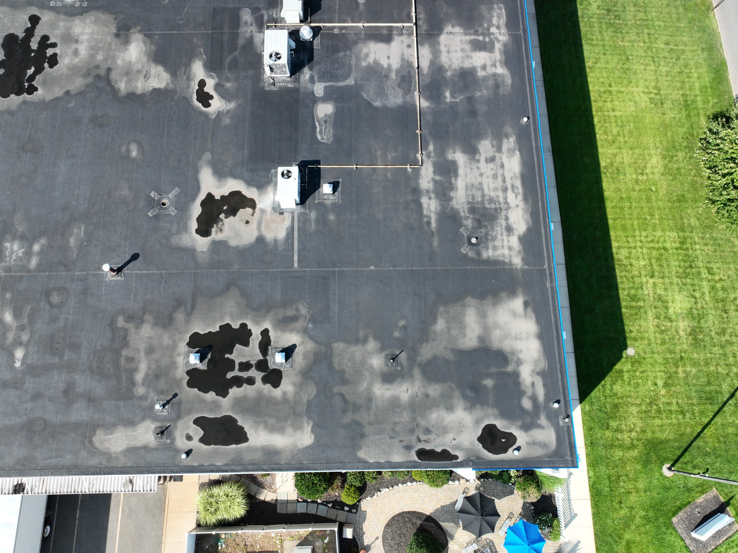 Aerial view of a flat rooftop with multiple patches of water damage and black stains, with some air conditioning units and small structures, next to a grassy area with trees and a backyard patio with umbrellas.