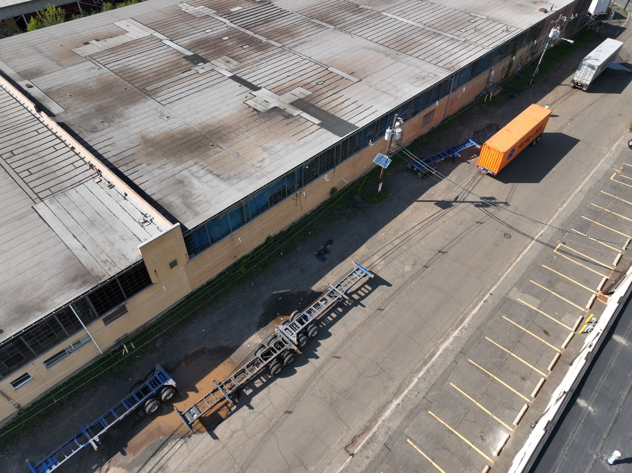 Aerial view of a large rooftop with patchwork metal roofing, adjacent empty parking lot, and a street with trucks and utility poles.