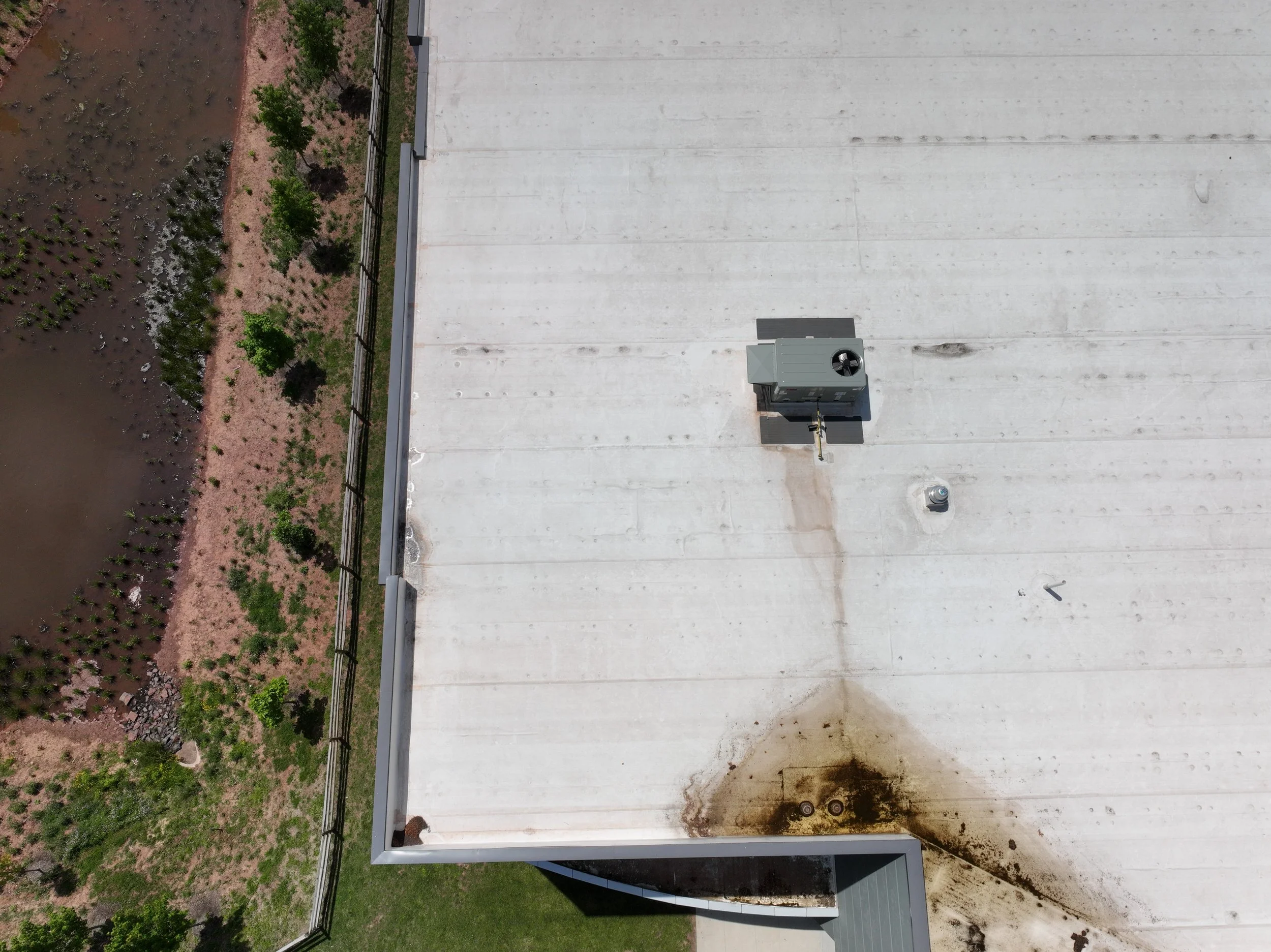 A flat white rooftop with install equipment, including cooling or heating units, with dirt and stains on the surface. The rooftop is bordered by a metal railing. Adjacent to the roof, there is landscape with small trees and bushes on reddish soil.