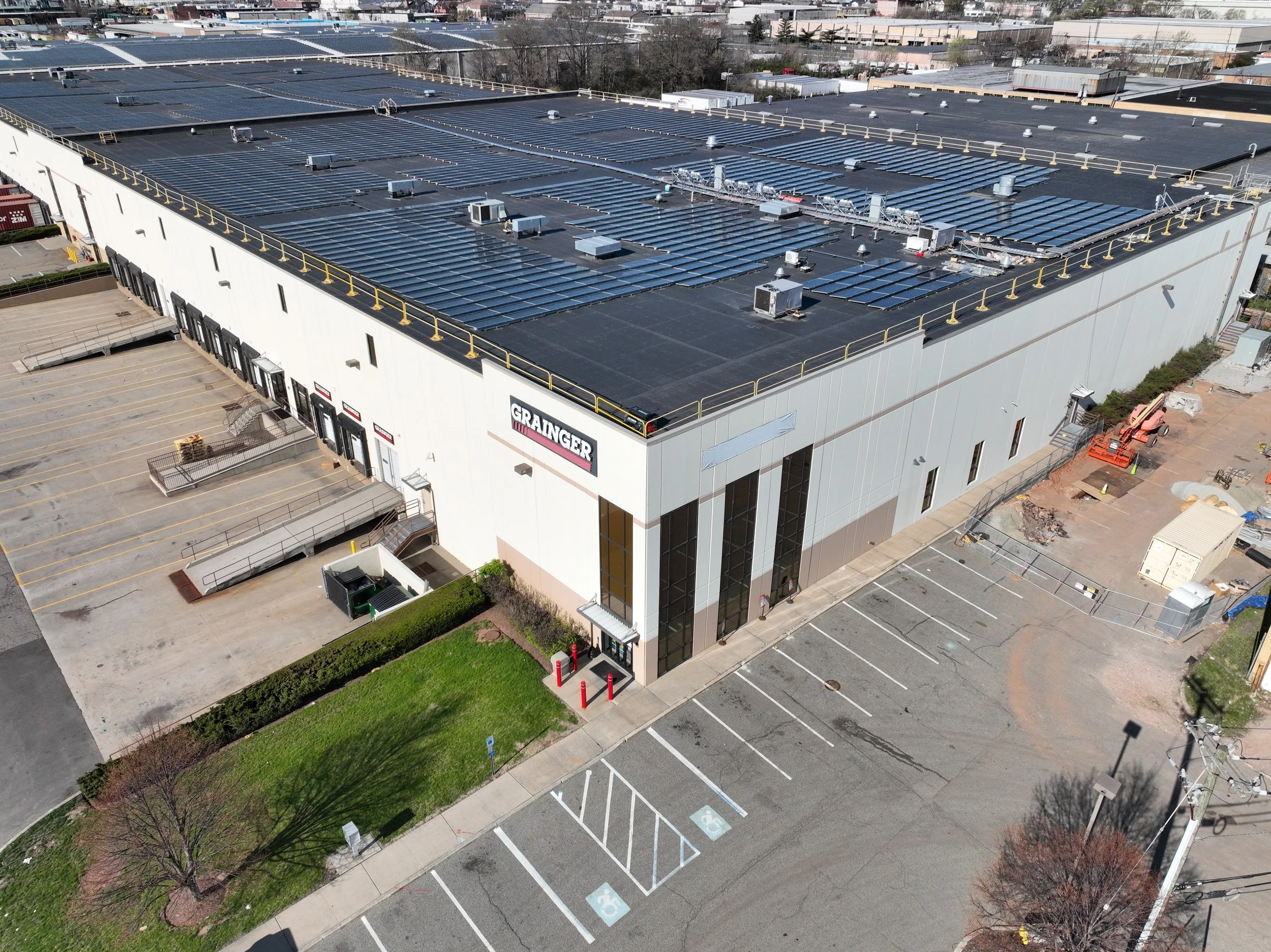 An aerial view of a large commercial building with a rooftop covered in solar panels. The building has a parking lot with designated spaces, including accessible parking, and some construction equipment nearby. The surrounding area consists of additional industrial buildings and parking lots.