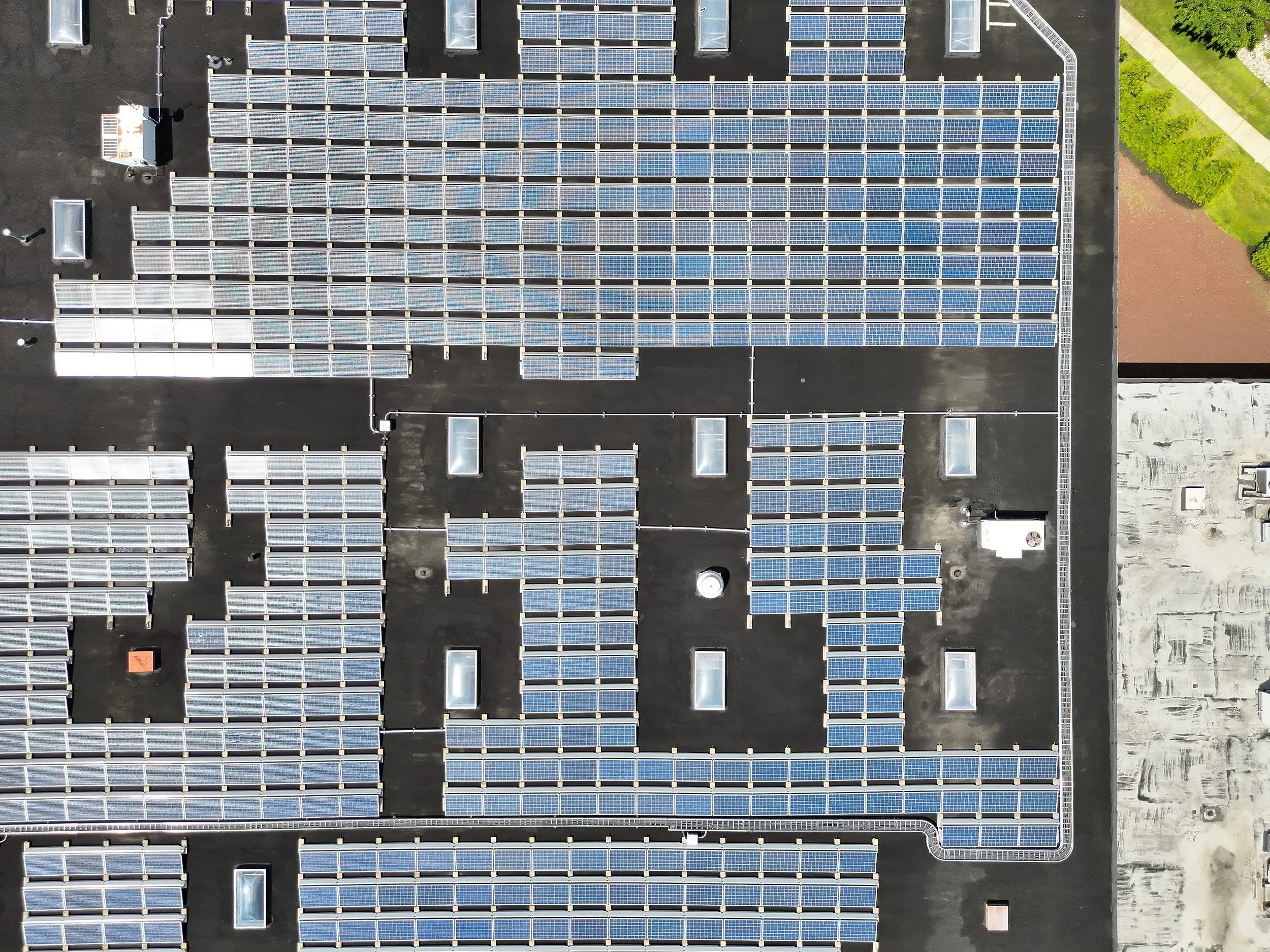 An aerial view of a large flat rooftop covered with solar panels, with some vents and a small enclosed structure, surrounded by some greenery and a sidewalk.
