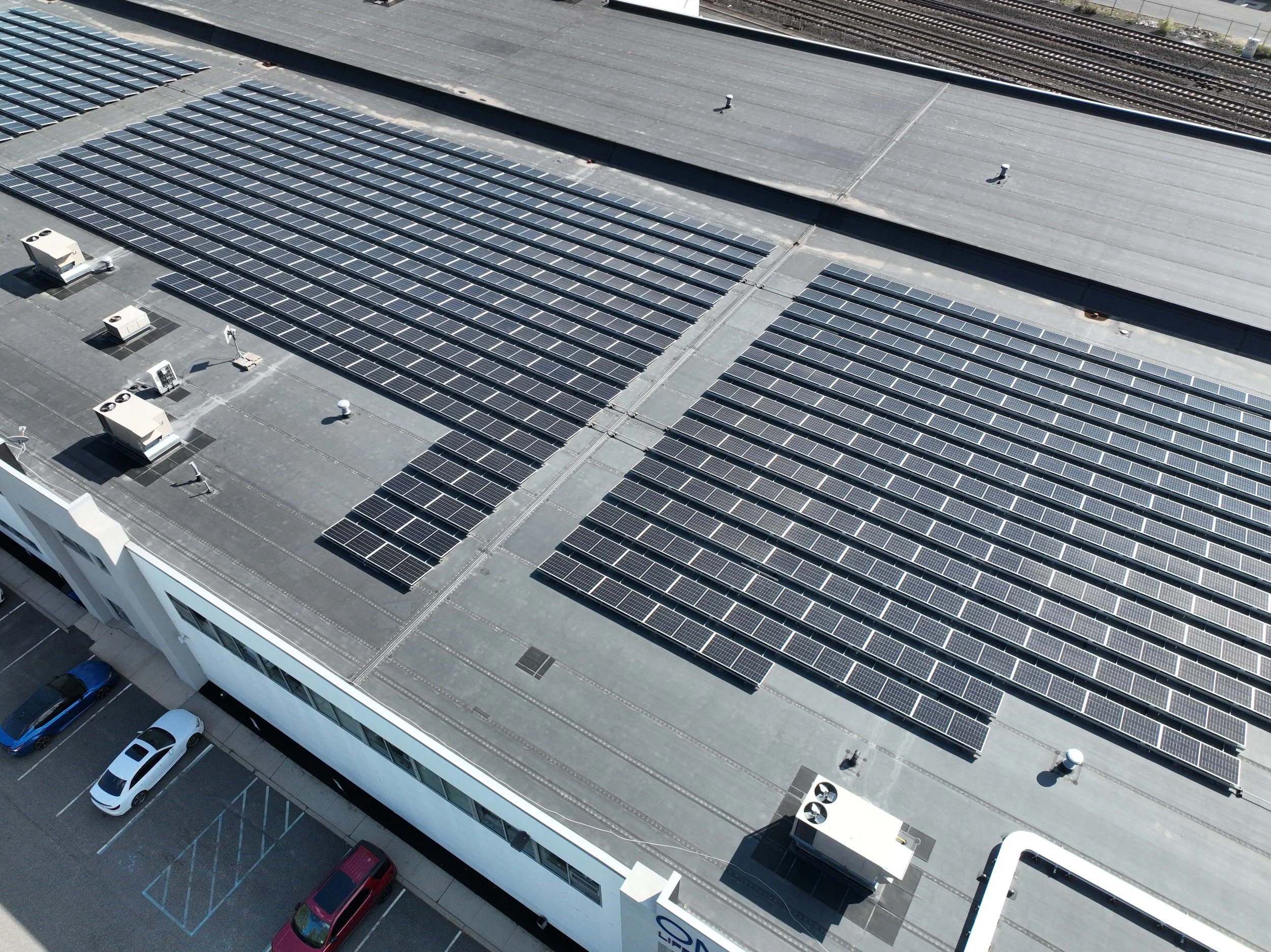 A building rooftop with rows of solar panels installed, visible vents, and air conditioning units, with parking below and train tracks in the background.