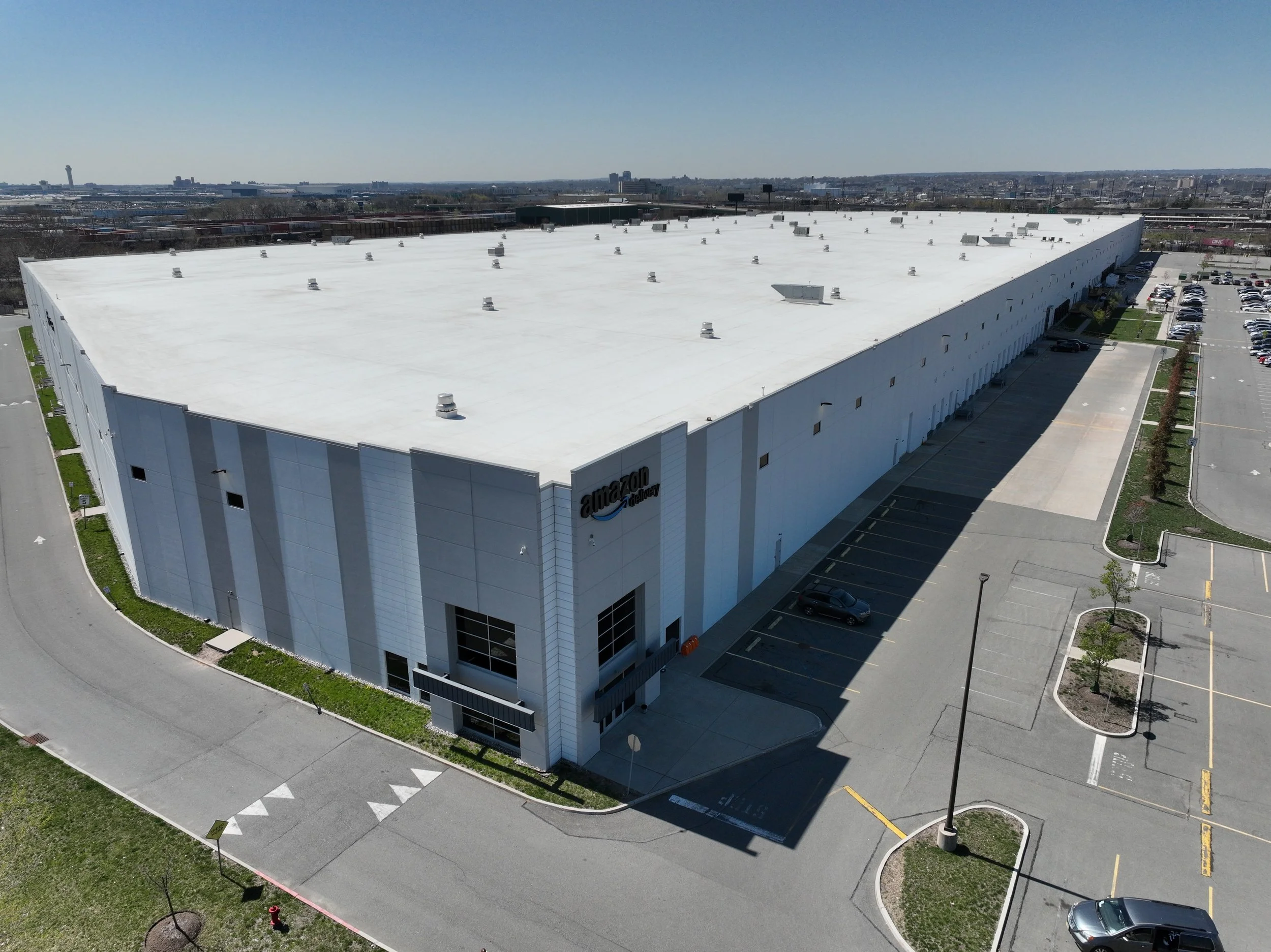 Aerial view of a large white commercial building with the Amazon logo, parking lot, and surrounding roads and cityscape in the background.