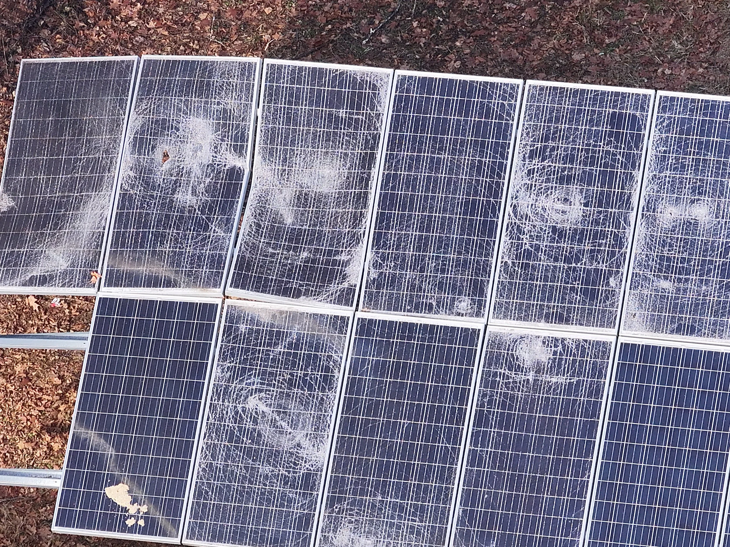 Multiple broken solar panels lying on the ground among dry leaves and soil.
