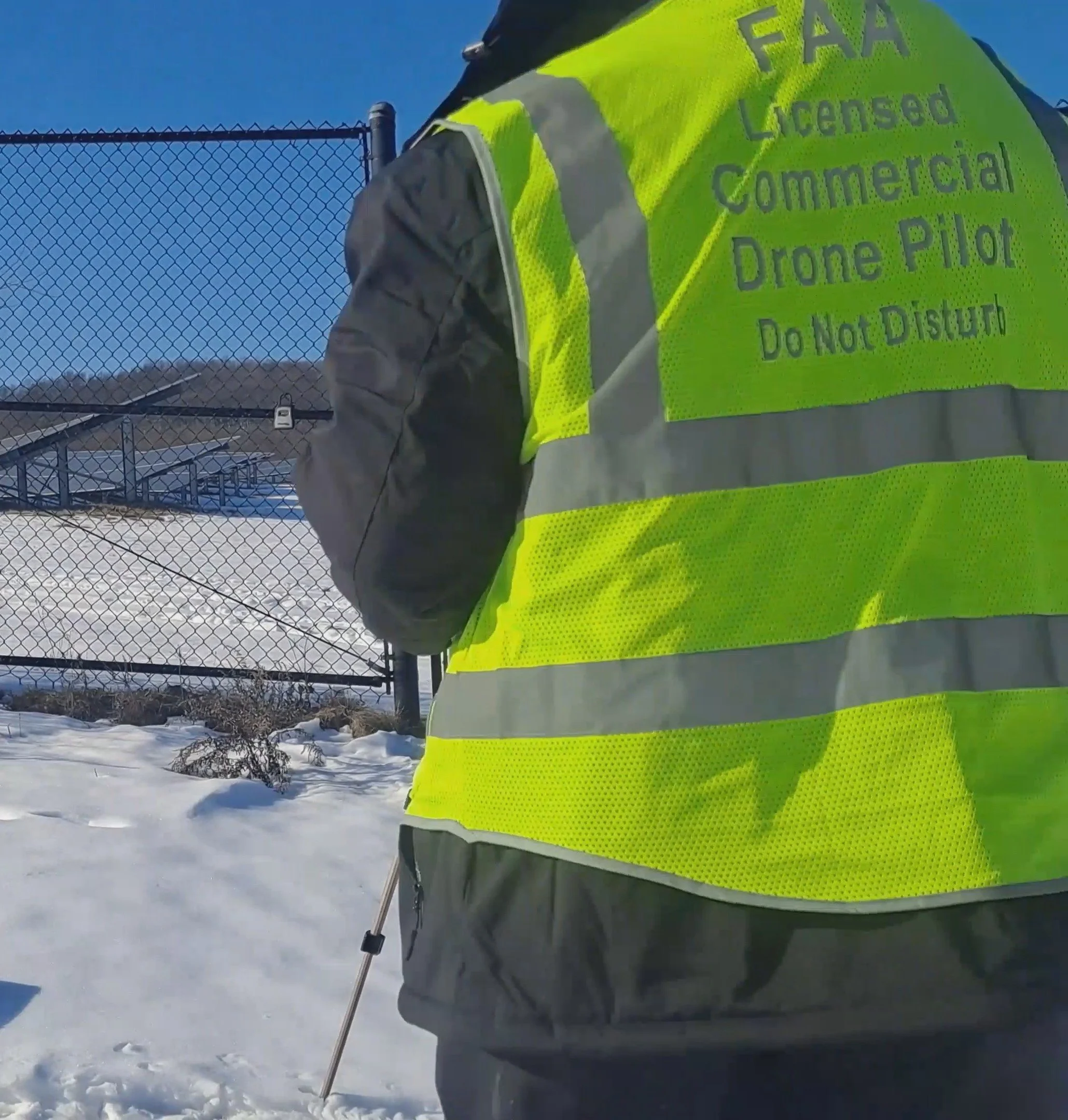 A person wearing a bright yellow safety vest with the words 'FAA Licensed Commercial Drone Pilot Do Not Disturb' printed on the back, standing outdoors in a snowy area near a chain-link fence under a clear blue sky.