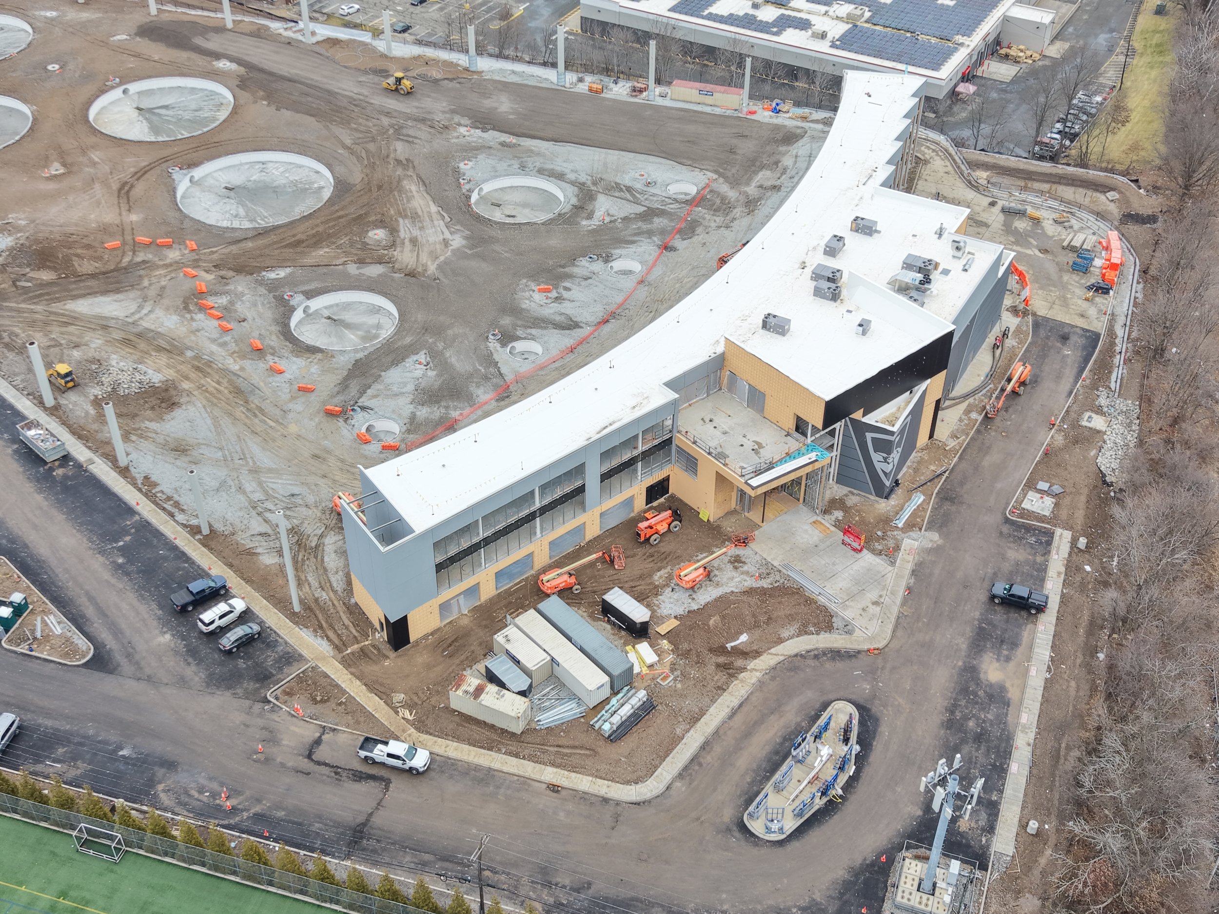 An aerial view of a construction site featuring a partially constructed building with a white roof, several large circular excavations with concrete borders, construction vehicles, and equipment, with a parking lot and trees surrounding the area.