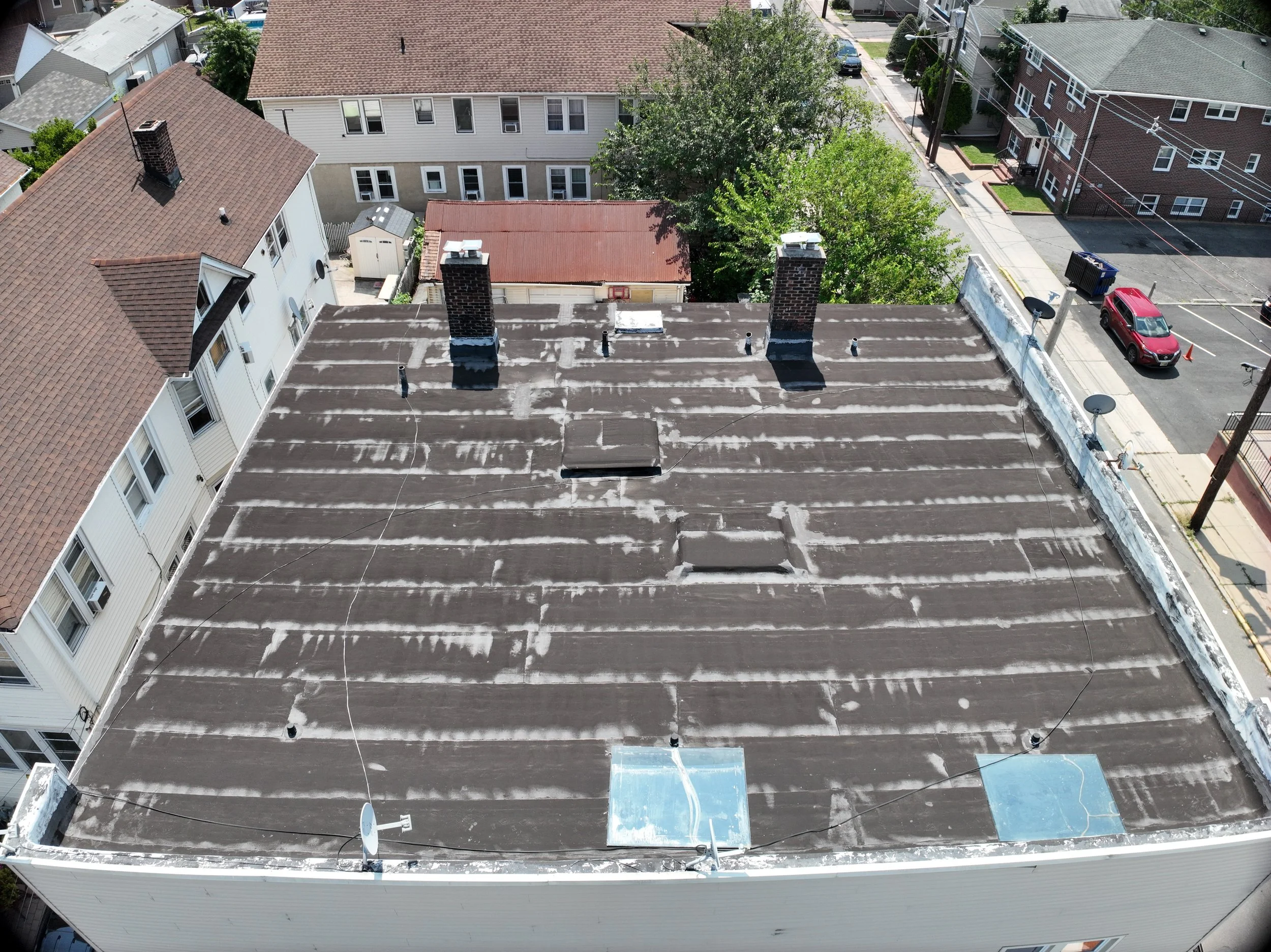 An aerial view of rooftops with various chimneys, satellite dishes, and vents, surrounded by residential buildings, trees, and a street with parked cars.