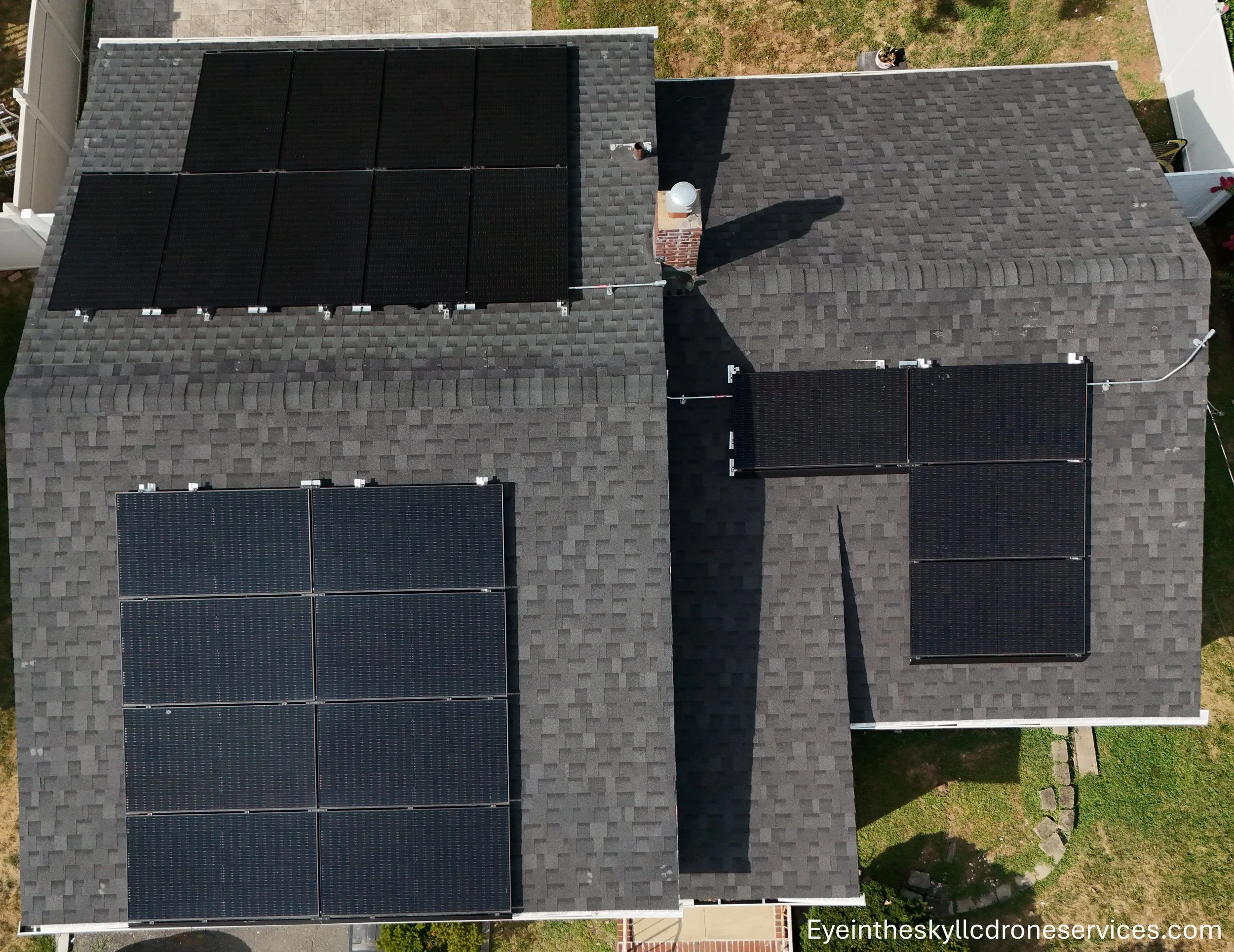 Aerial view of a house with solar panels on two sections of the roof, chimney, and a yard with some grass and pavement.