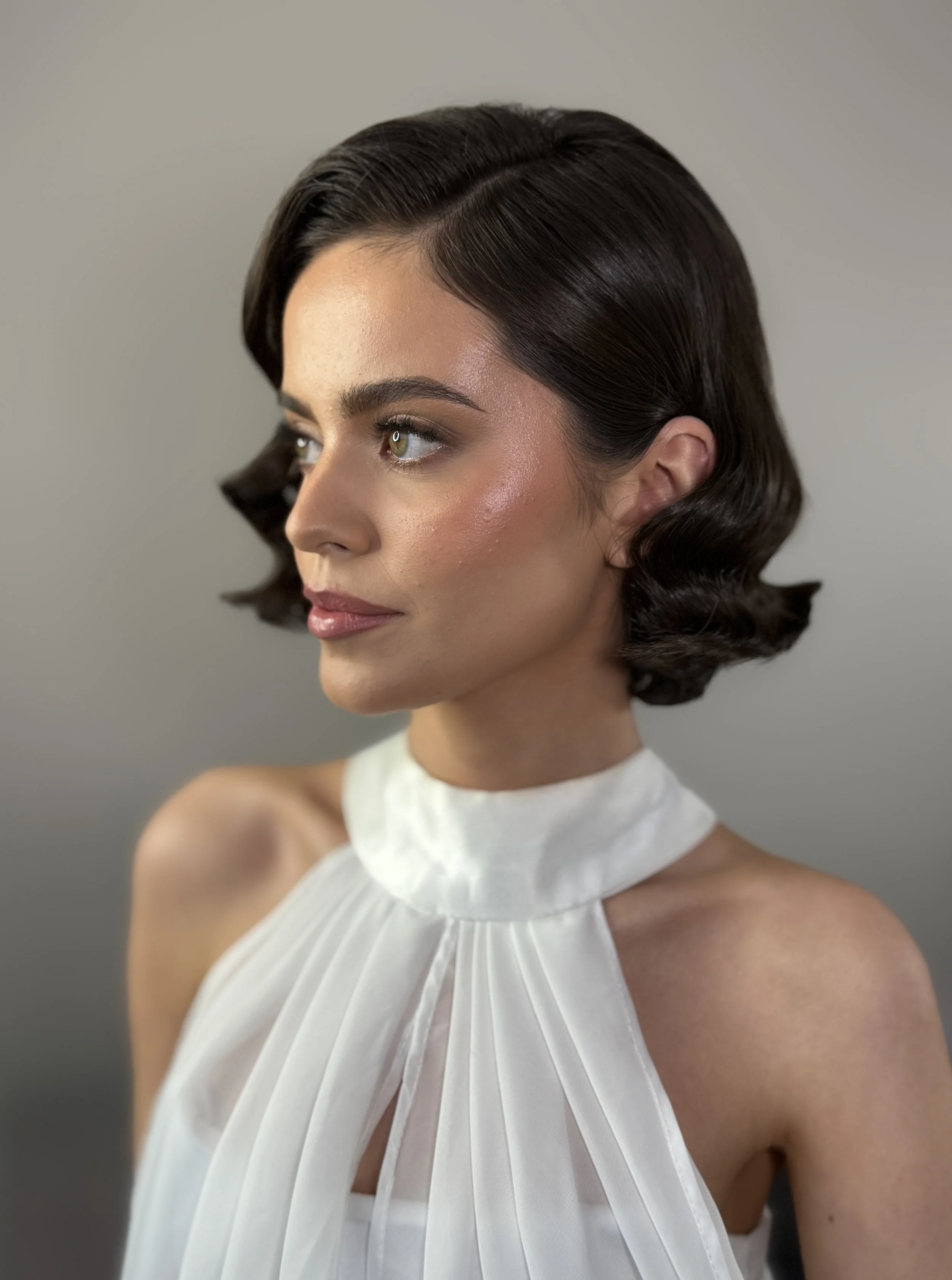 A woman with short, dark, wavy hair and olive green eyes, wearing a white, pleated, high-neck sleeveless top. She has makeup with natural tones, glossy lips, and a subtle blush. The background is plain and gray.