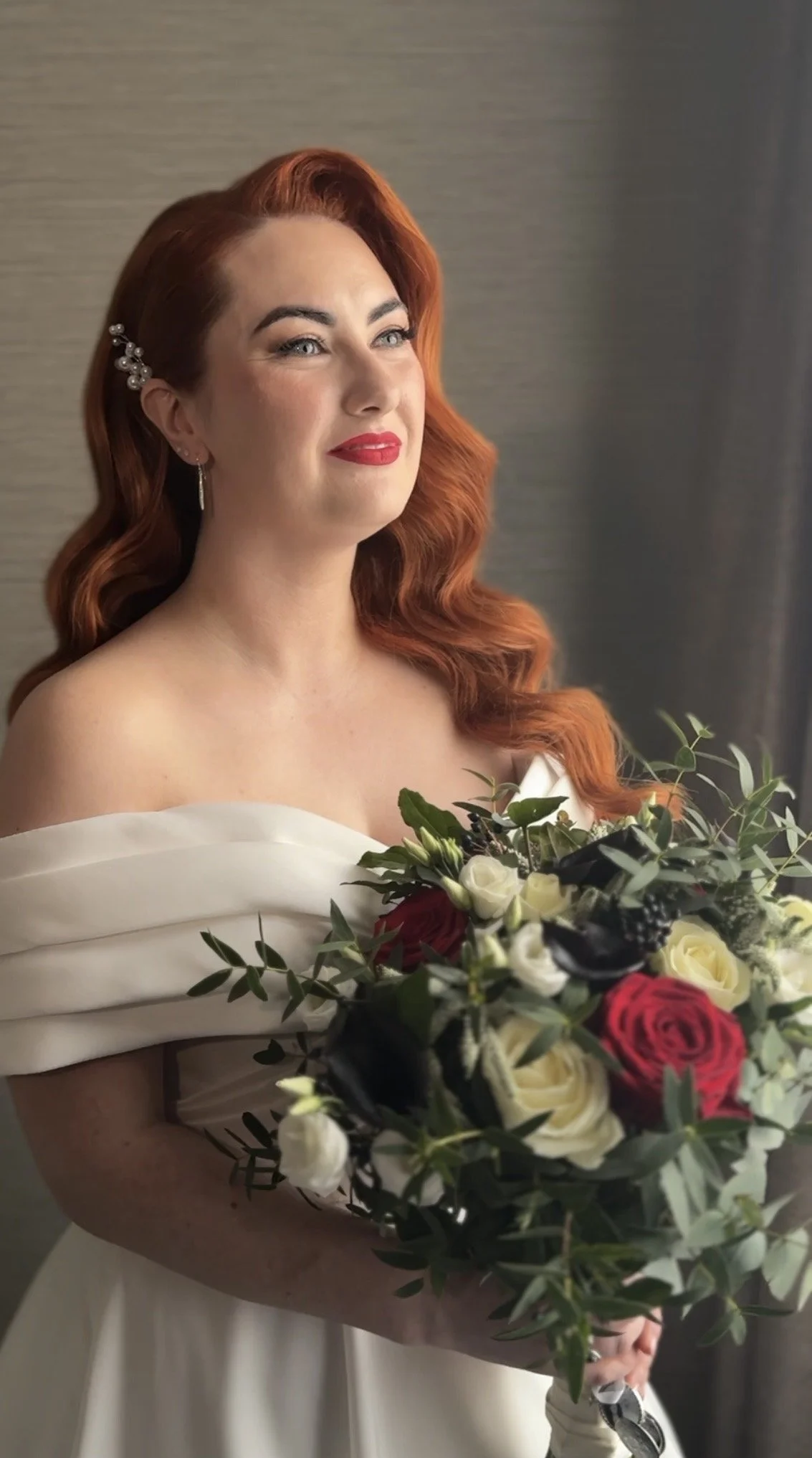 A woman with red hair in vintage waves, wearing pearl earrings and a white off-shoulder dress, holding a bouquet of red, white, and black roses, standing near a gray wall.