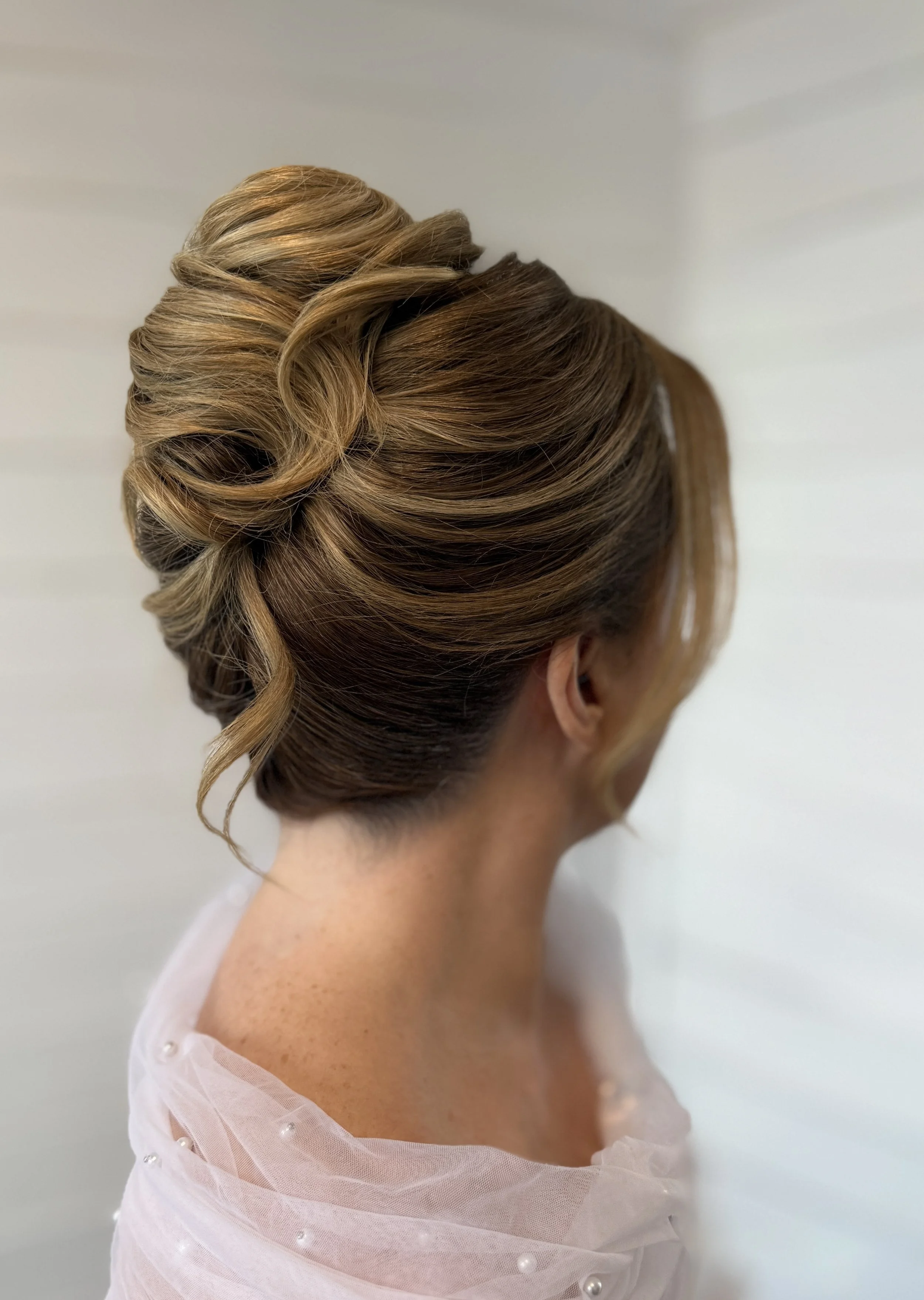 Elegant updo hairstyle with curled sections and highlights on a woman with fair skin, wearing a sheer pink garment with pearl embellishments.