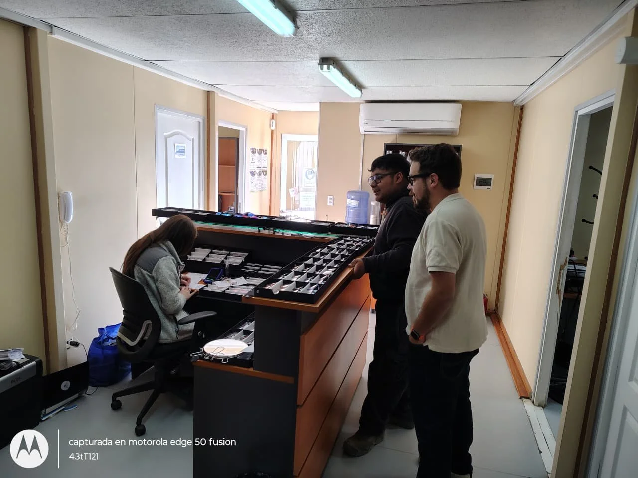 Dos hombres conversando en un mostrador con diversos objetos y una mujer sentada trabajando en una oficina con muebles y equipo tecnológico.