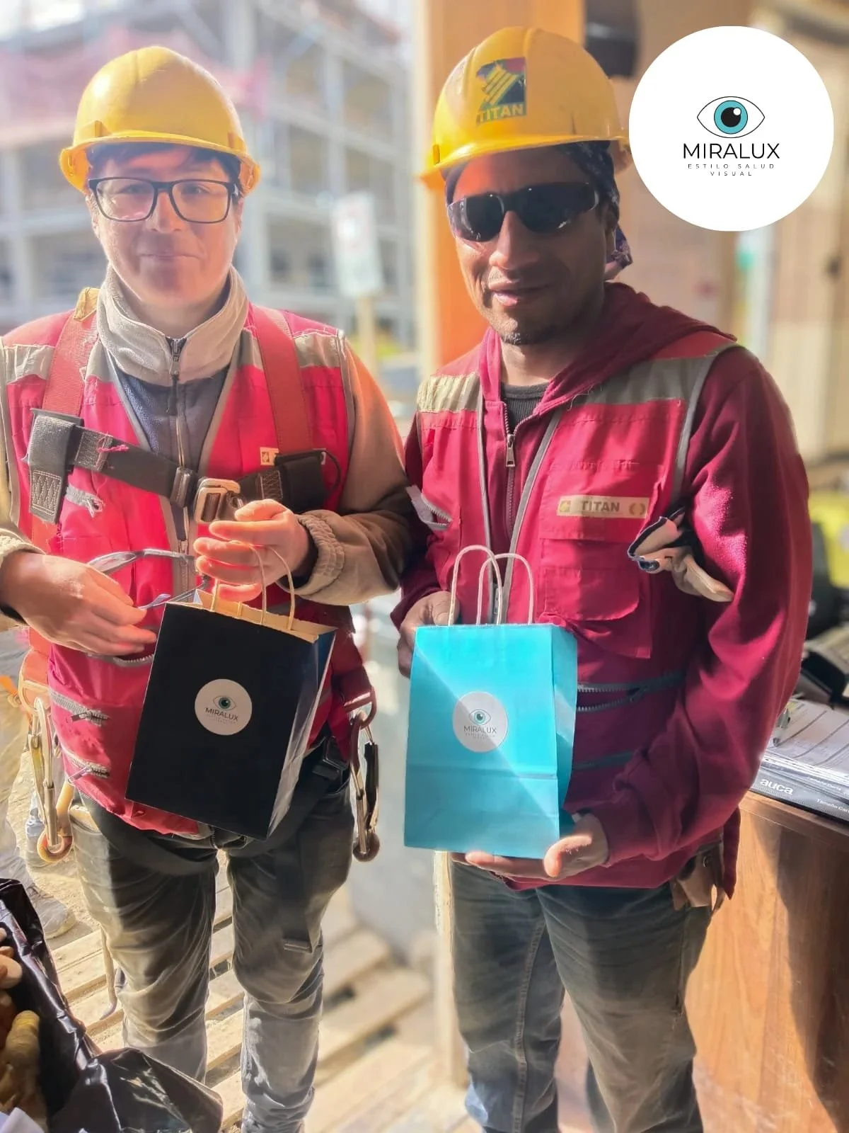 Dos hombres con casco y ropa de trabajo sosteniendo bolsas de regalo en un ambiente de construcción o taller.