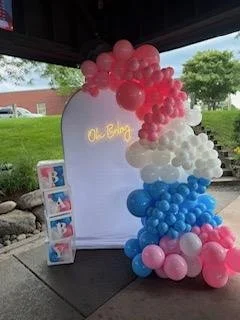Balloon arch with pink, white, blue, and light pink balloons, shaped like a rainbow, set up outdoors under a pavilion. A small stand with flyers is nearby.