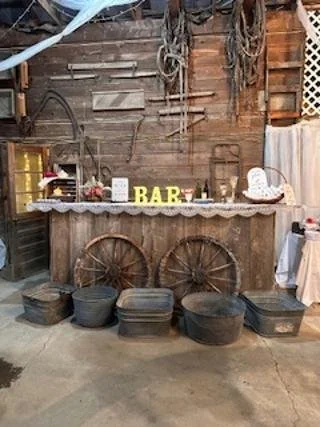 Rustic outdoor bar with wooden wheels, buckets on the ground, and vintage tools hanging on a wooden wall.