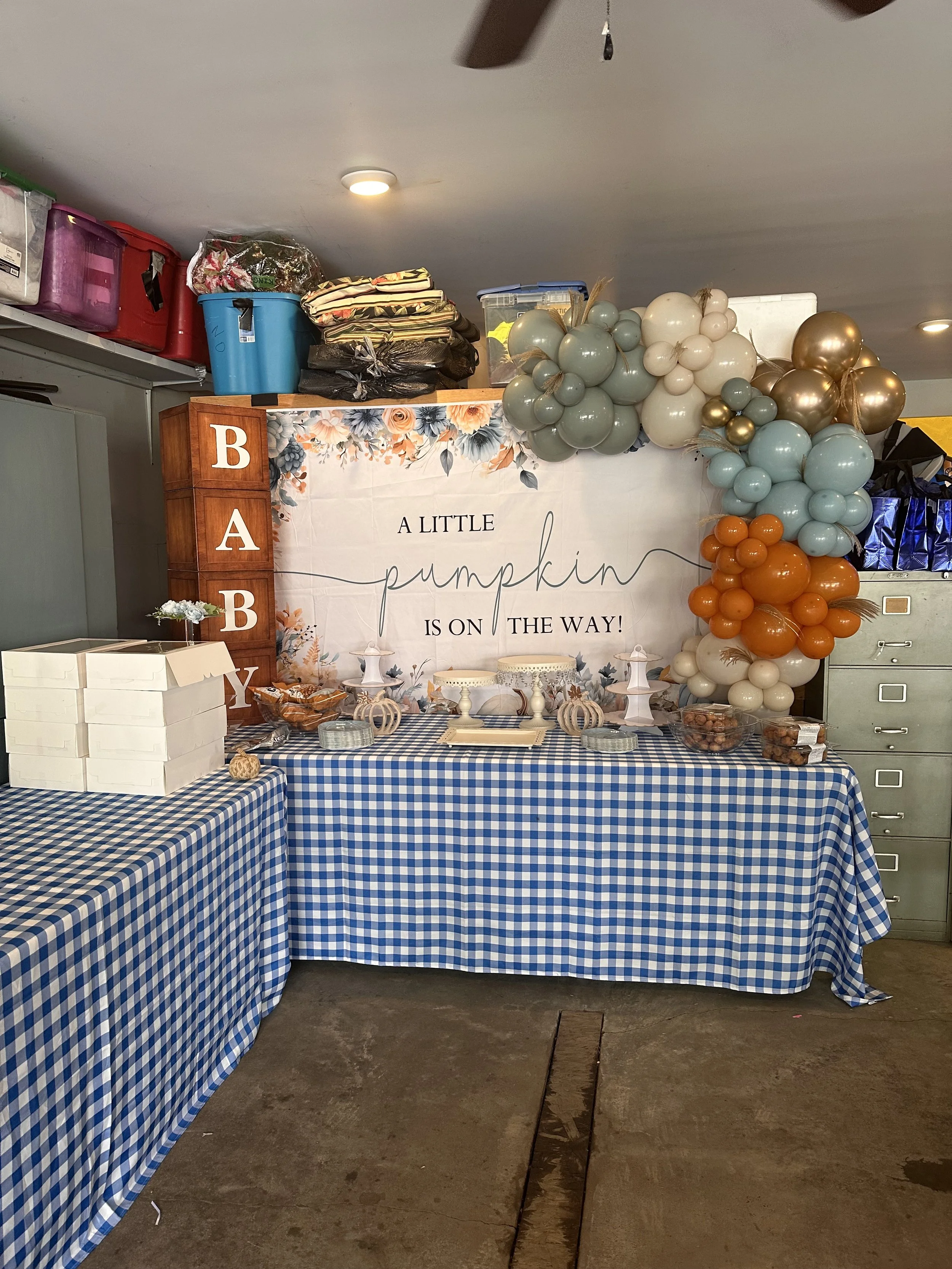 Party setup with a blue gingham tablecloth, a large sign that reads "A Little Pumpkin Is On The Way!", and balloon arrangements in gray, cream, orange, and gold. There are small cakes and snacks on the table, with boxes for guests to take home treats.