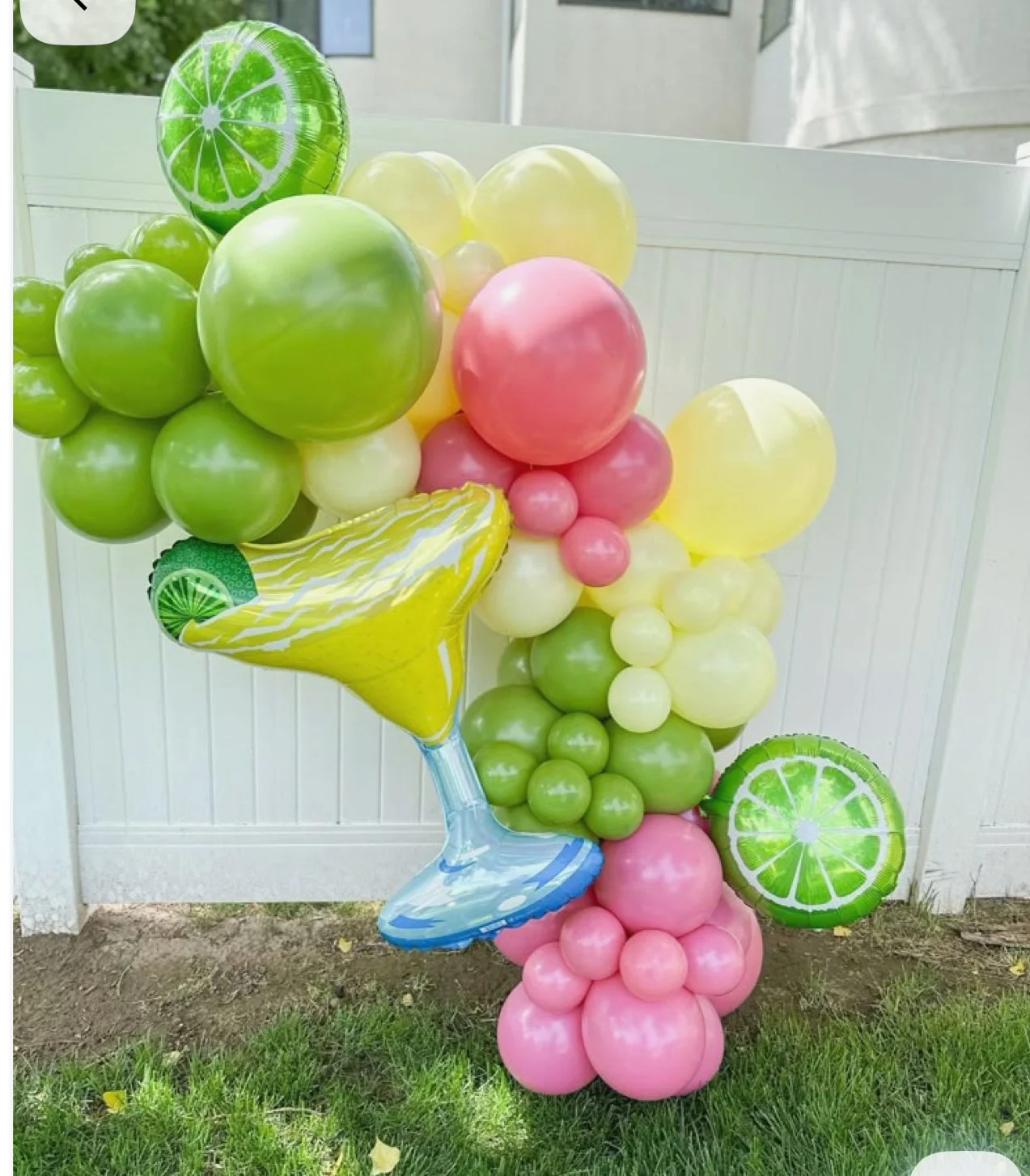 Colorful balloon arrangement with green, pink, yellow, and lime-themed balloons, set outdoors on grass with a white fence in the background.