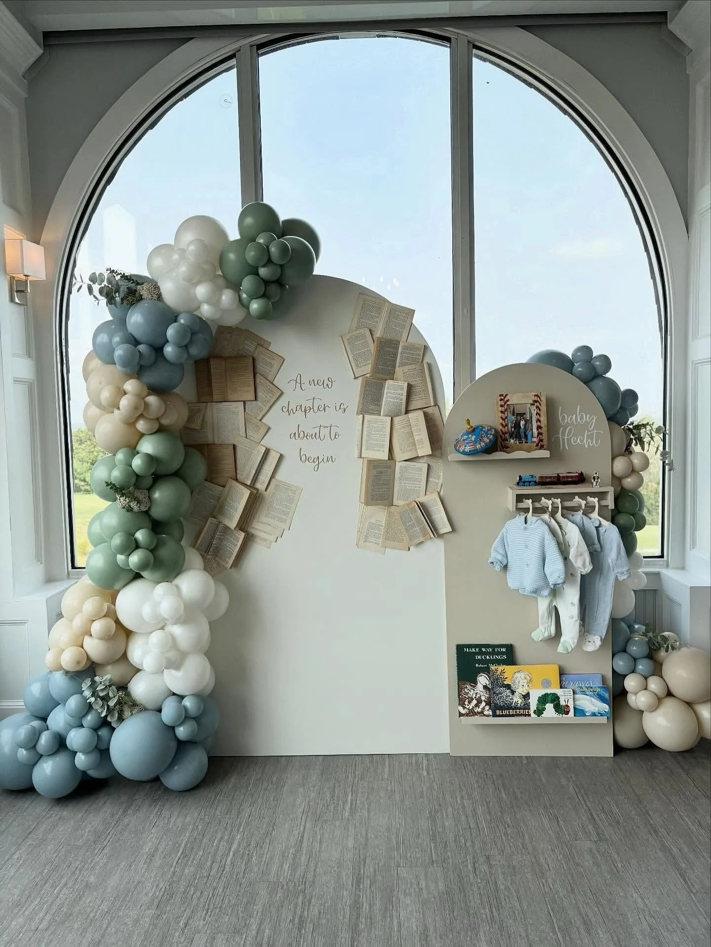 Decorative baby shower display with balloons, open books, and a hanging clothing rack for baby clothes, set against a large window and with a sign reading 'A new chapter is about to begin.'