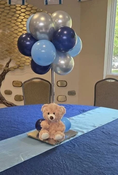 A teddy bear sitting on a tray with blue and dark blue balloons attached to a bunch of balloons behind it, on a table covered with a blue tablecloth in a decorated room.
