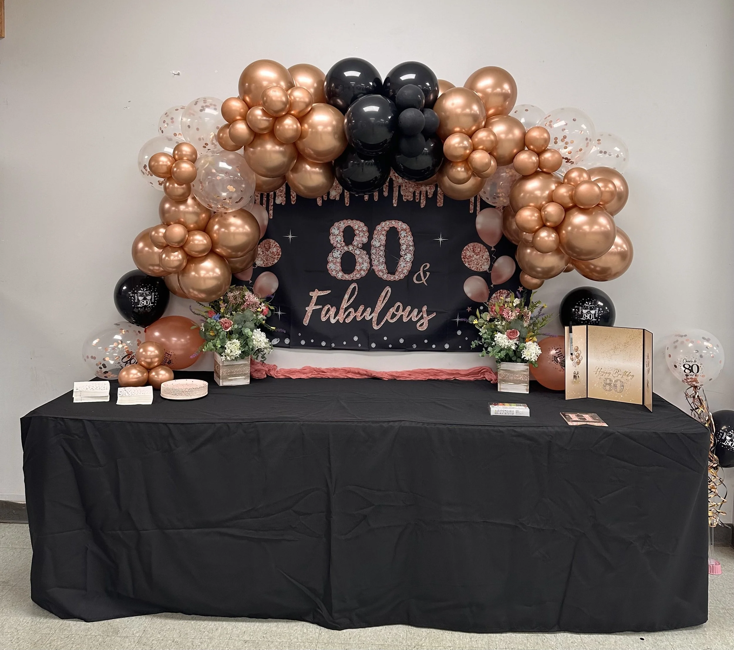 Decorative backdrop for an 80th birthday celebration with a black banner, pink and black balloons, floral arrangements, and birthday cards on a table.