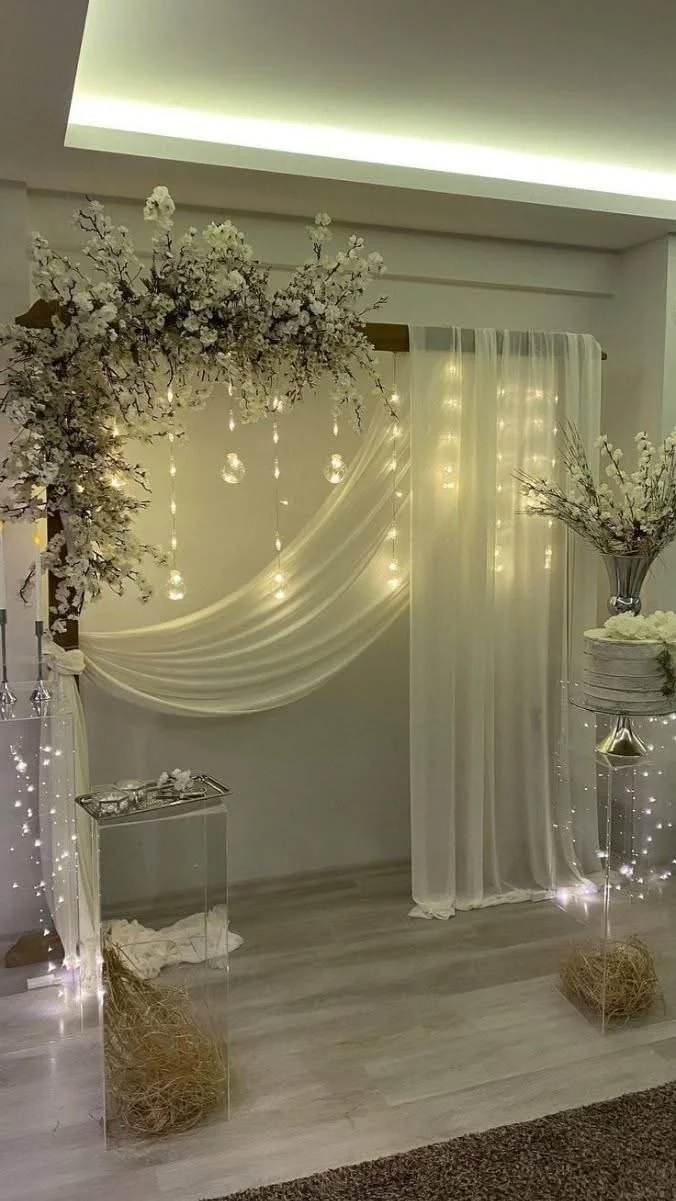 Elegant indoor scene with white floral arrangements, sheer curtains, hanging fairy lights, and a soft ceiling light, creating a cozy and decorative atmosphere.