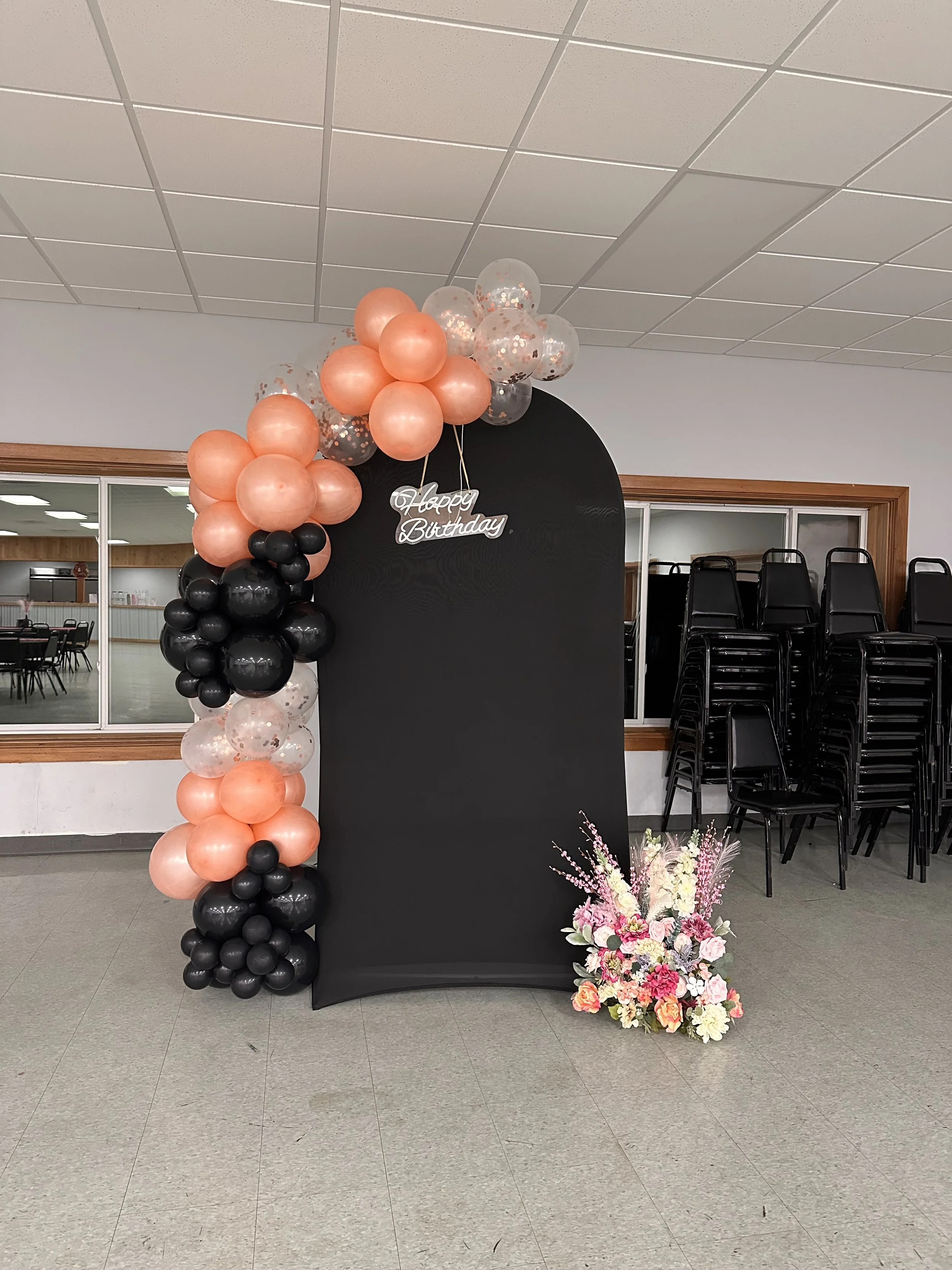 Decorative backdrop with a black backdrop, pink, black, and clear balloons arranged on the left side, and a bouquet of pink, white, and peach flowers on the floor to the right. A silver birthday sign hangs on the backdrop reading 'Happy Birthday'.