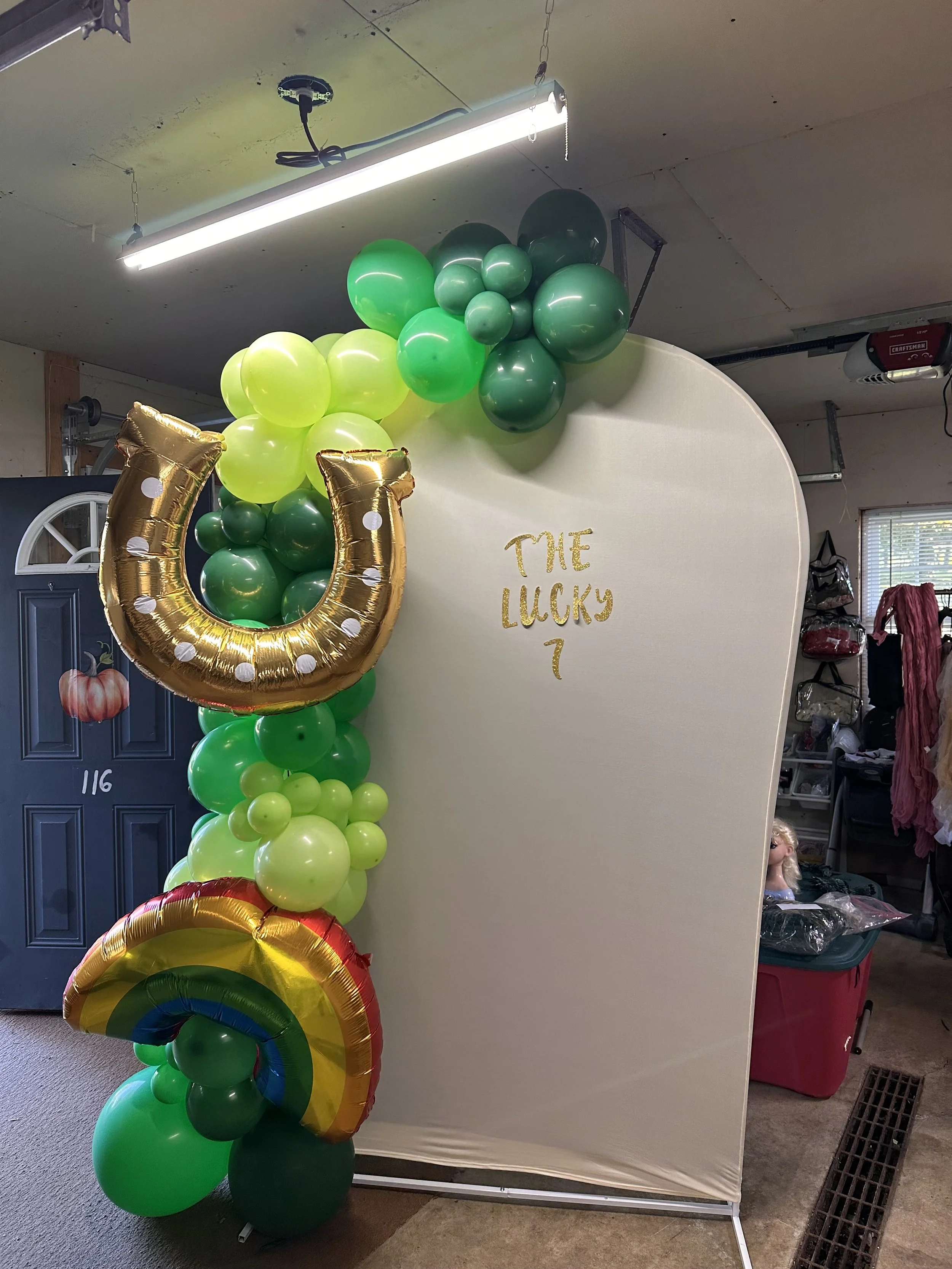 Decorative balloon arch with green, yellow, and rainbow balloons, including a gold horseshoe balloon and a rainbow-shaped balloon, standing beside a fabric backdrop with golden text that reads "THE LUCKY 7".