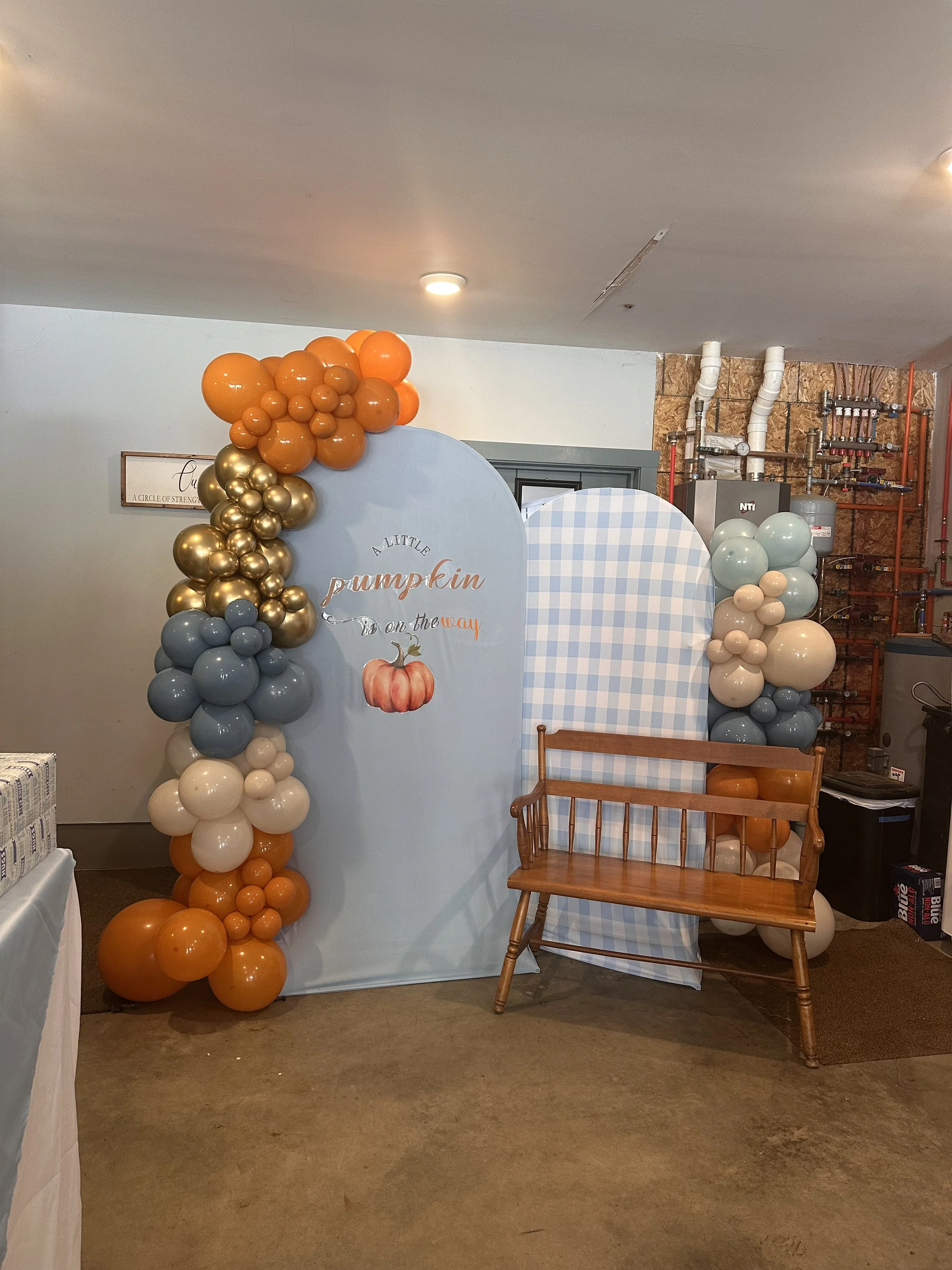 Decorative backdrop with balloon arch in orange, gold, blue, beige, and white, featuring a sign reading 'A Little Pumpkin is on the Way' with a pumpkin illustration, and a wooden bench in front.