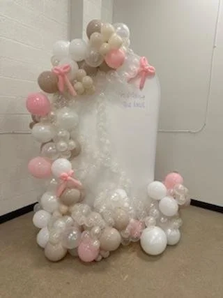 Decorative balloon arch with pink, white, beige, and peach balloons, some with pink ribbons, around a white backdrop.