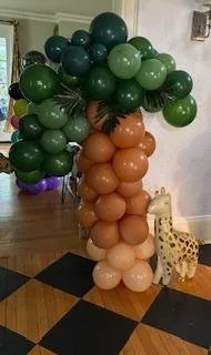 Balloon sculpture of a palm tree with green and tan balloons, and a yellow giraffe toy nearby, on a checkered floor.