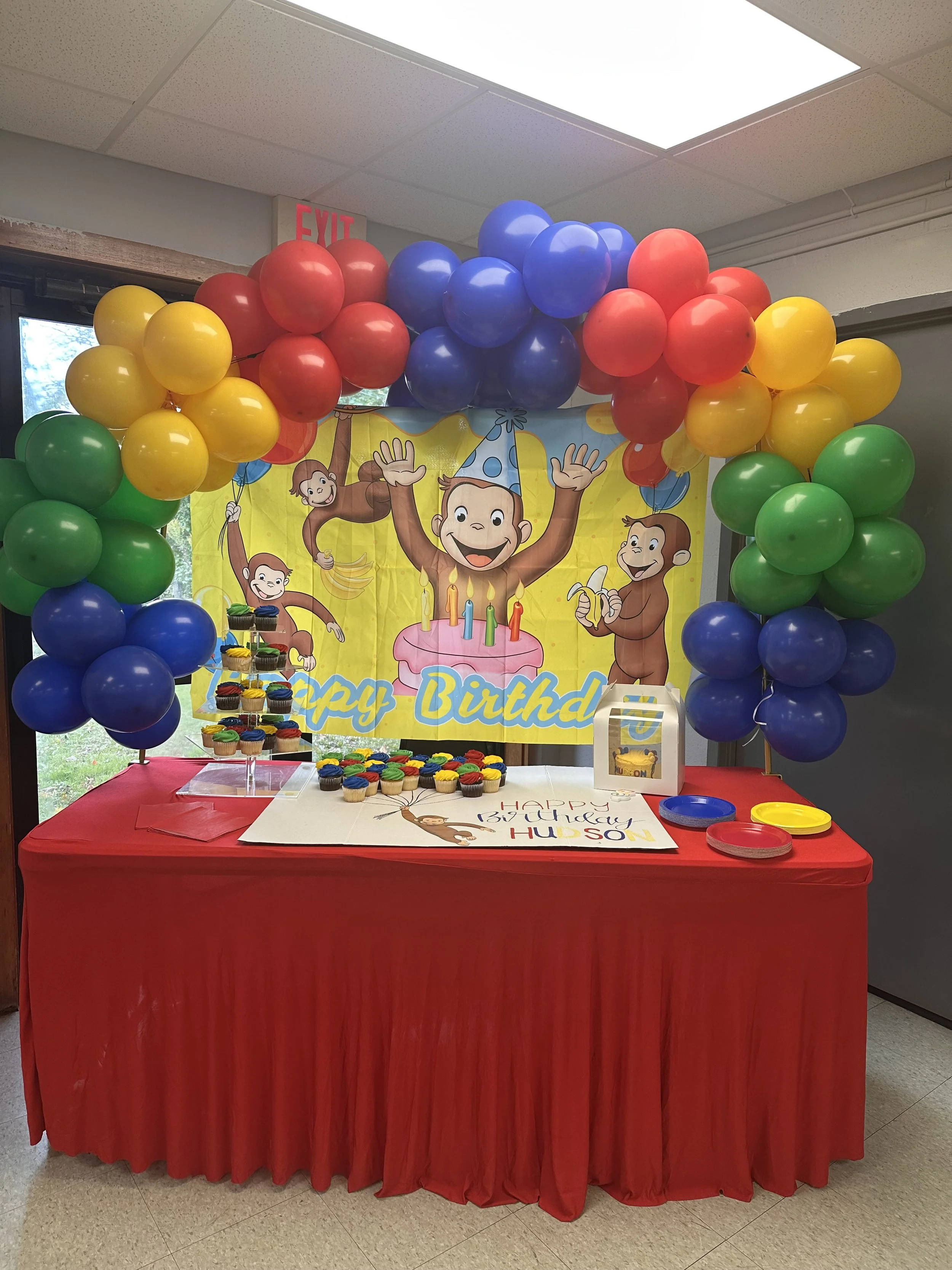 Colorful birthday party table with cupcakes, balloons, and a poster of monkeys celebrating around a birthday cake.