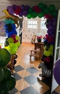Balloon arch with various colored balloons including purple, red, green, yellow, and blue decorating an indoor space with a wooden table and chairs near a window.