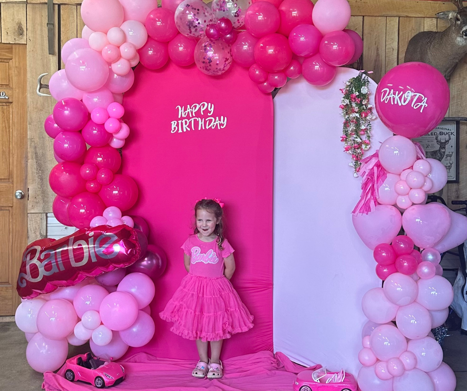 Little girl in pink dress standing in front of a pink and white birthday backdrop decorated with pink balloons, a large pink Barbie balloon, and a pink toy car at a Barbie-themed birthday celebration.